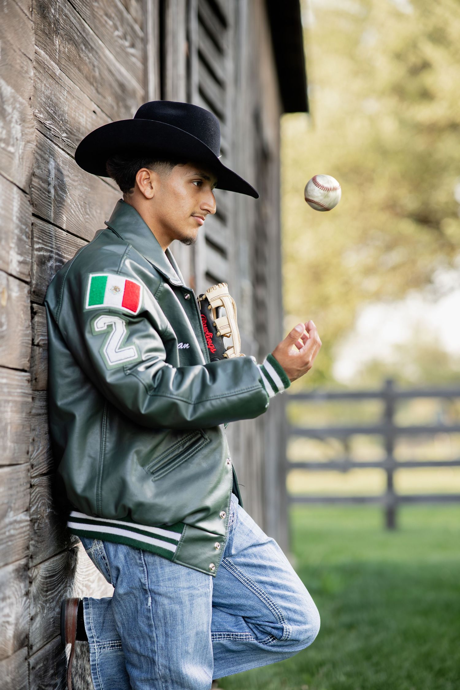 Young man in green varsity jacket and cowboy hat tossing baseball against rustic wooden wall.