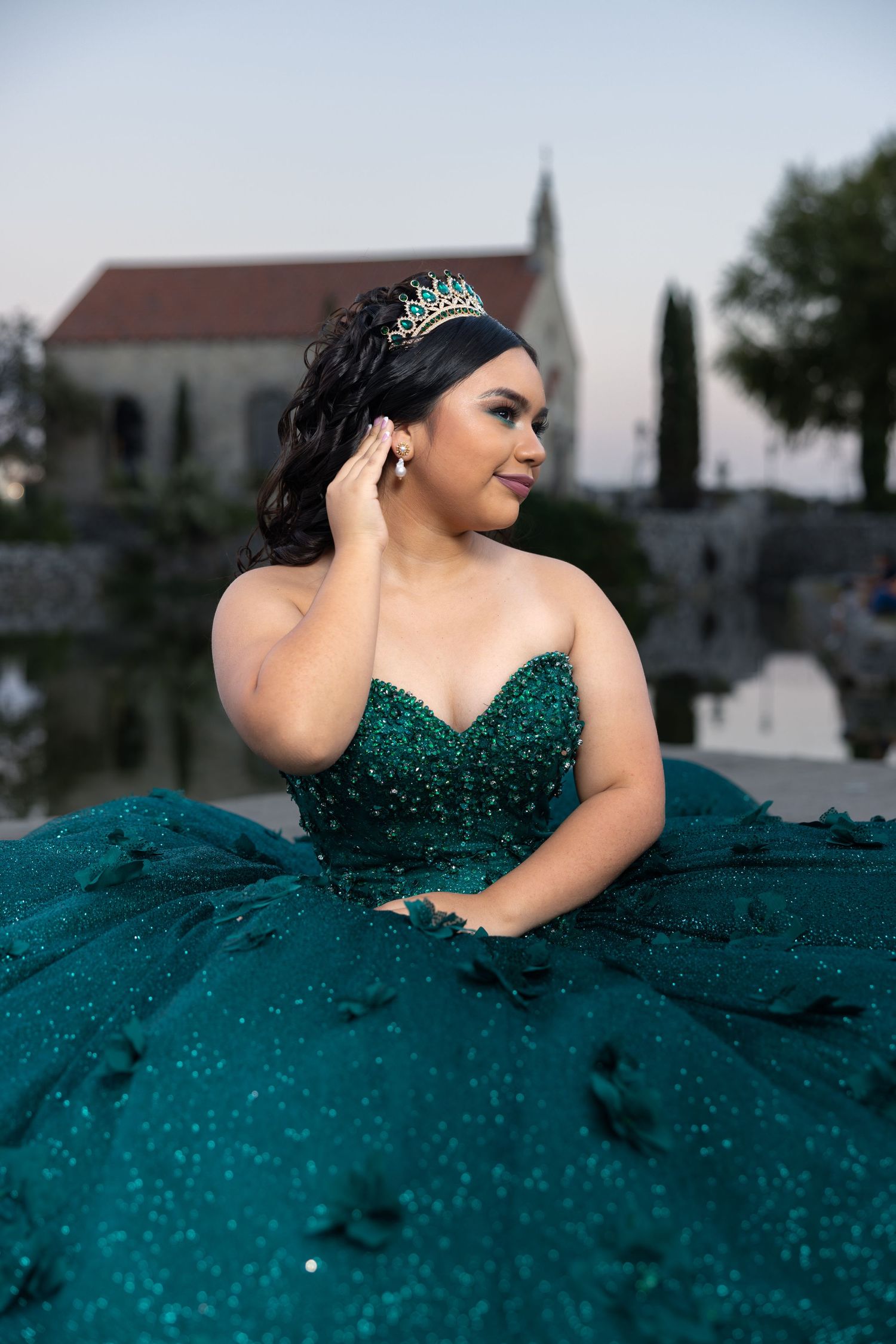 Young woman in an emerald green beaded quinceañera ball gown wearing a tiara outdoors.