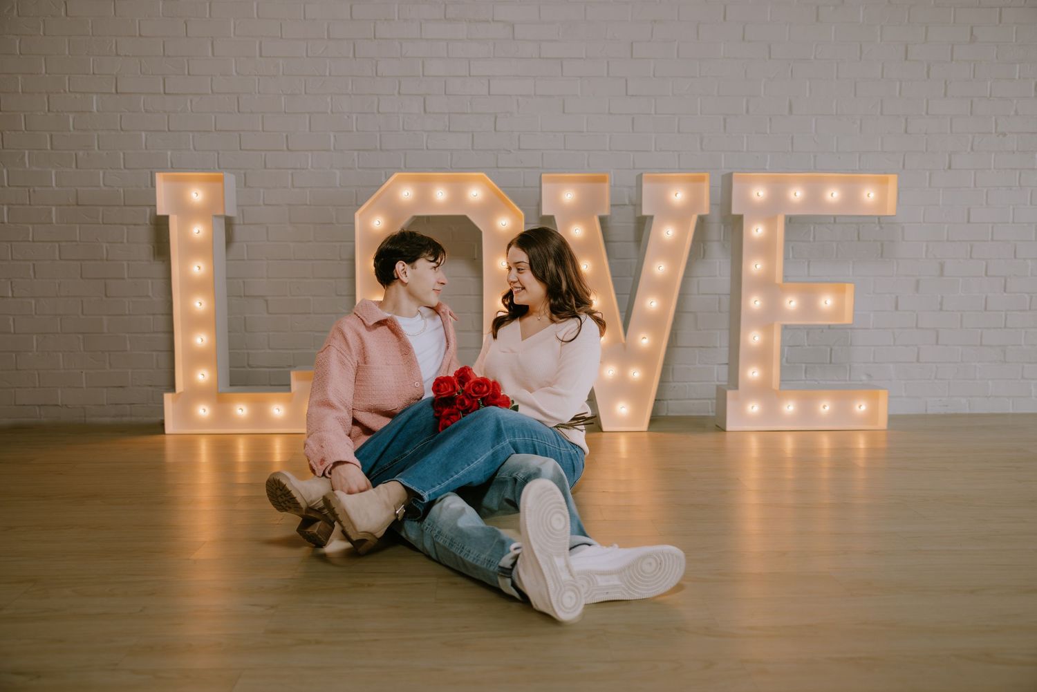 Romantic couple sitting on floor in front of illuminated LOVE marquee letters holding red roses.