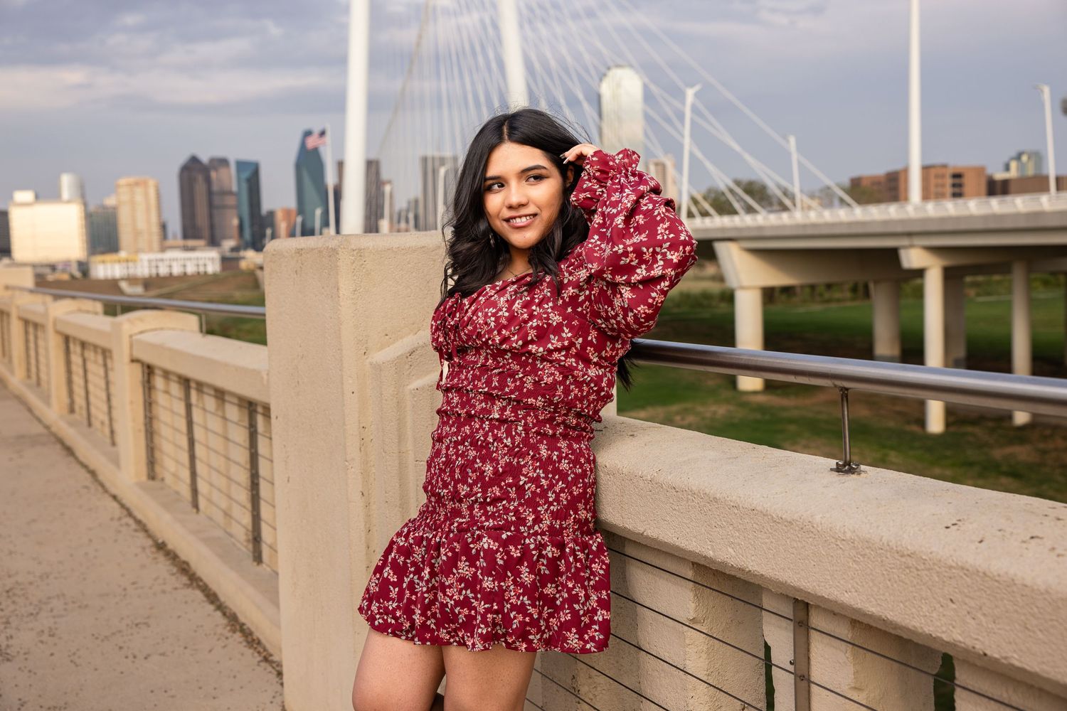 A young woman in a red floral mini dress poses on a bridge with a city skyline backdrop.