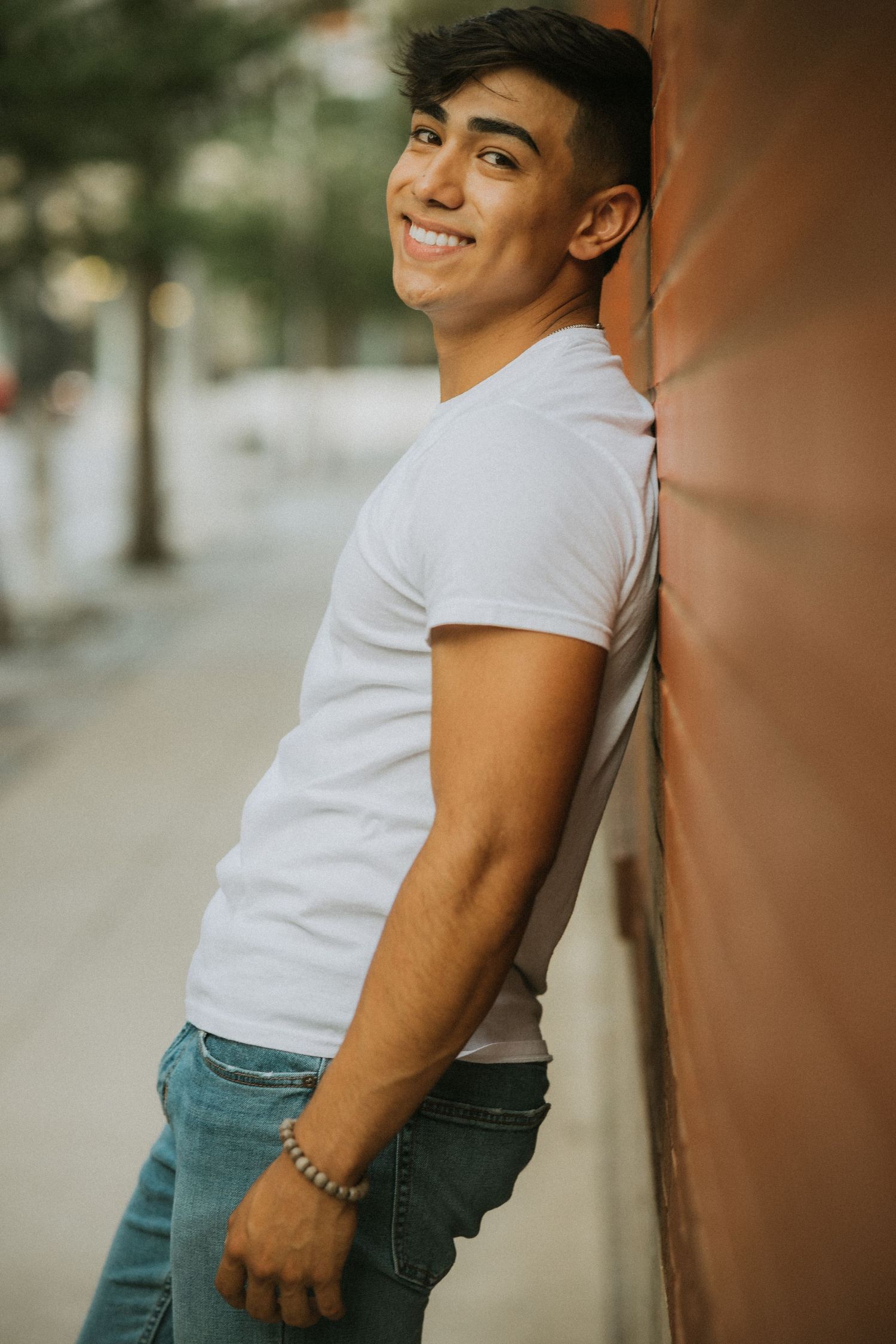 Smiling young man in white t-shirt leans against brown wall in outdoor urban portrait.