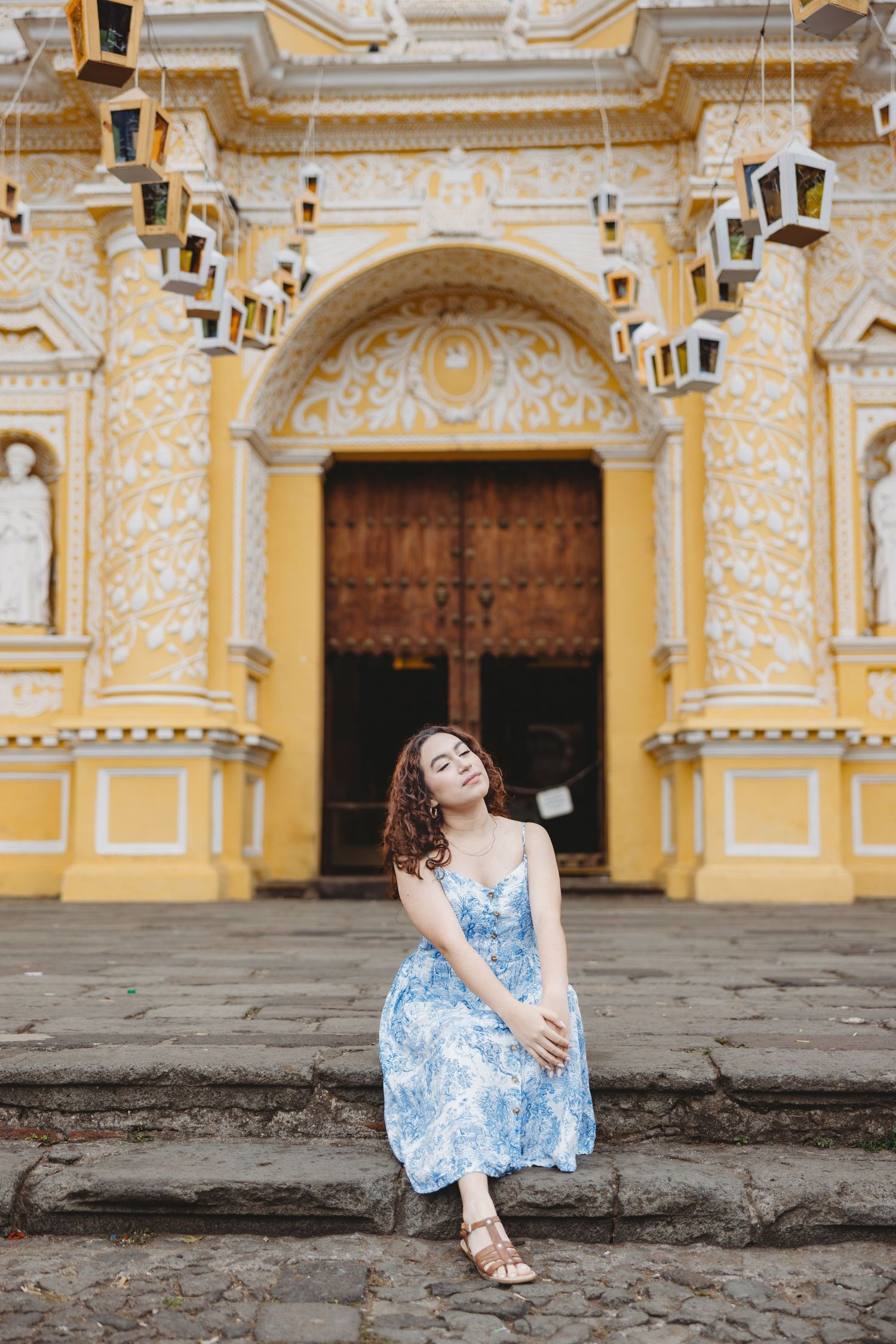 Young woman in blue floral dress sitting on steps of ornate yellow church.
