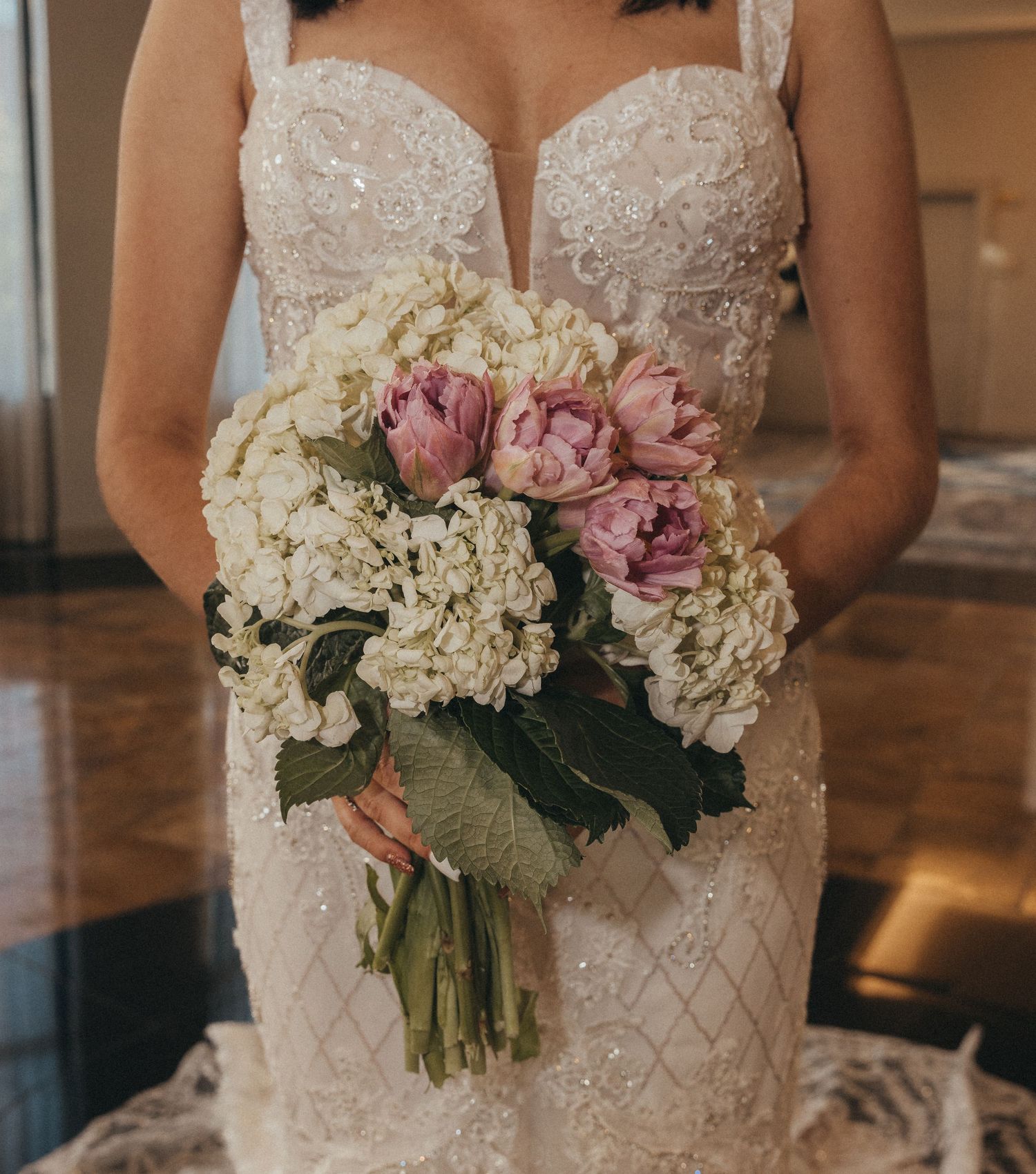 Bride holds elegant bouquet of white hydrangeas and pink tulips against beaded lace wedding gown.