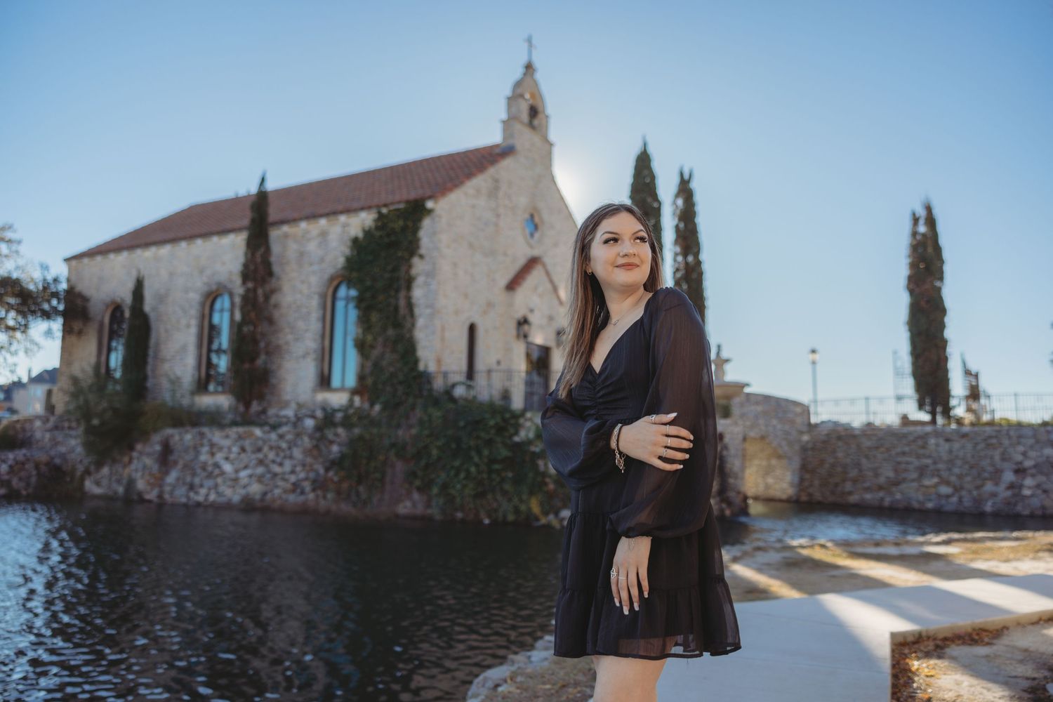 Woman in black dress posing outdoors near historic stone church and water feature.