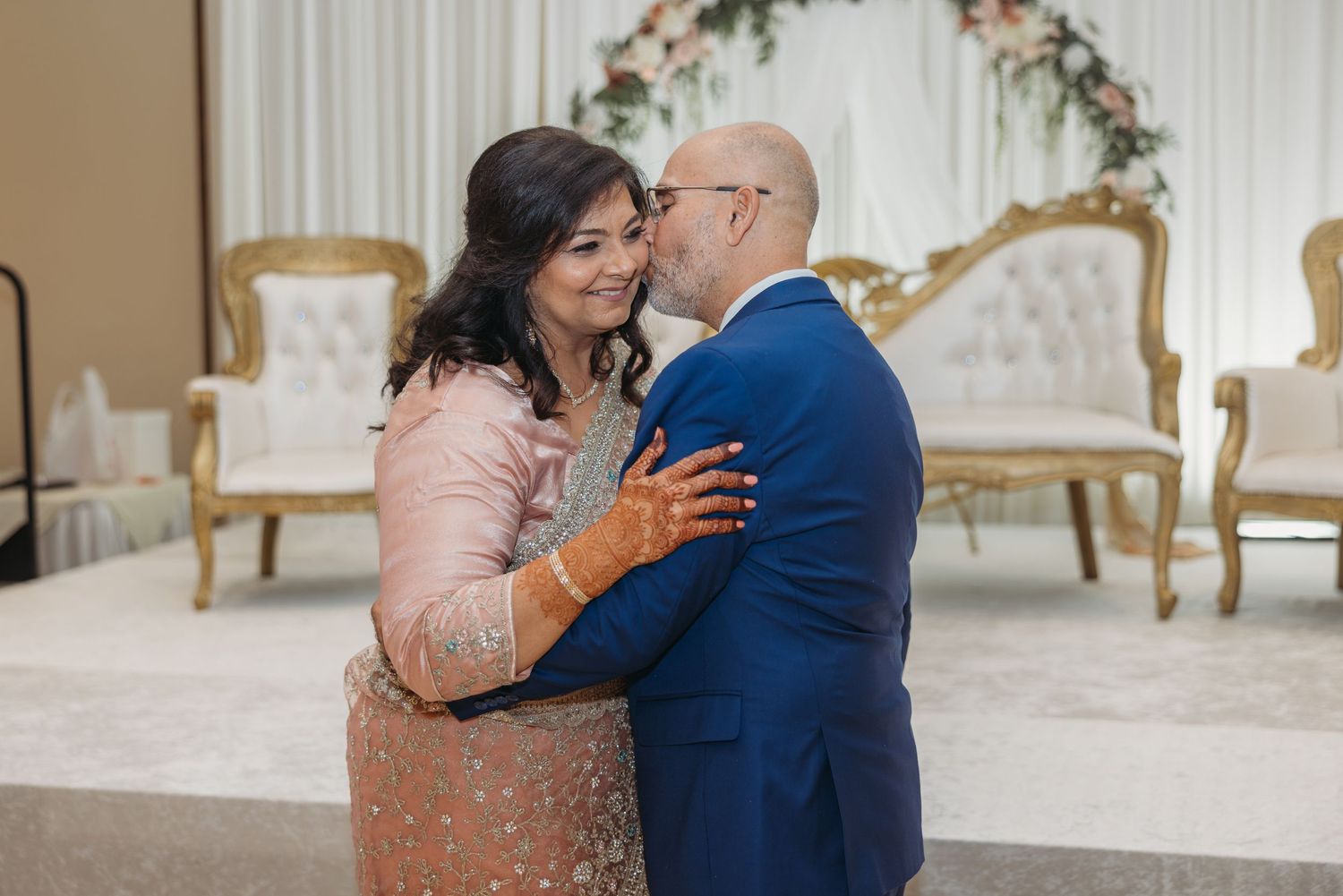Couple shares first dance embrace in elegant ballroom with floral arch and gold chairs.
