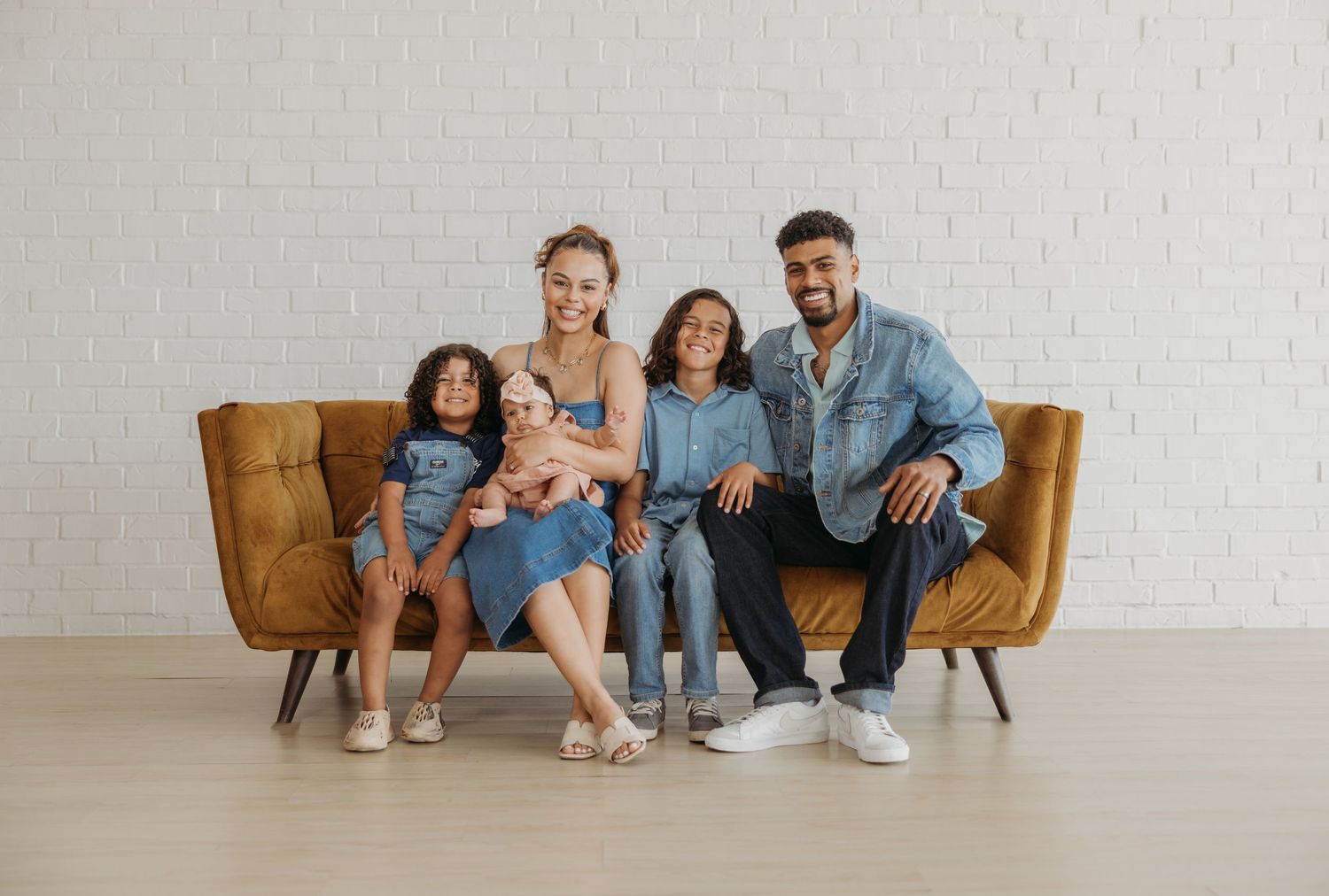 Smiling family of five in casual denim outfits posing together on a mustard yellow couch indoors.