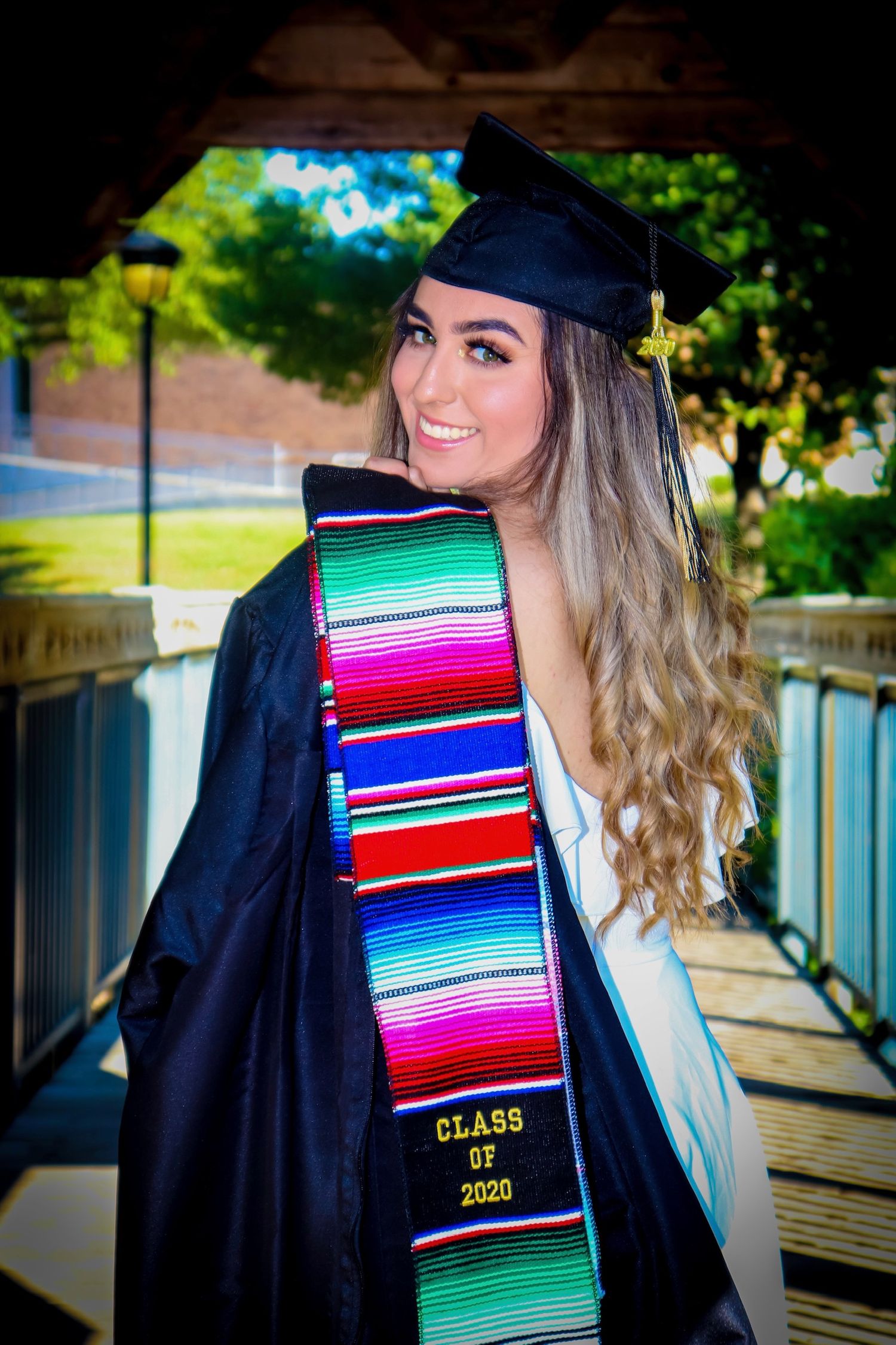 Graduate in black cap and gown with colorful Class of 2020 sash smiles over shoulder outdoors.