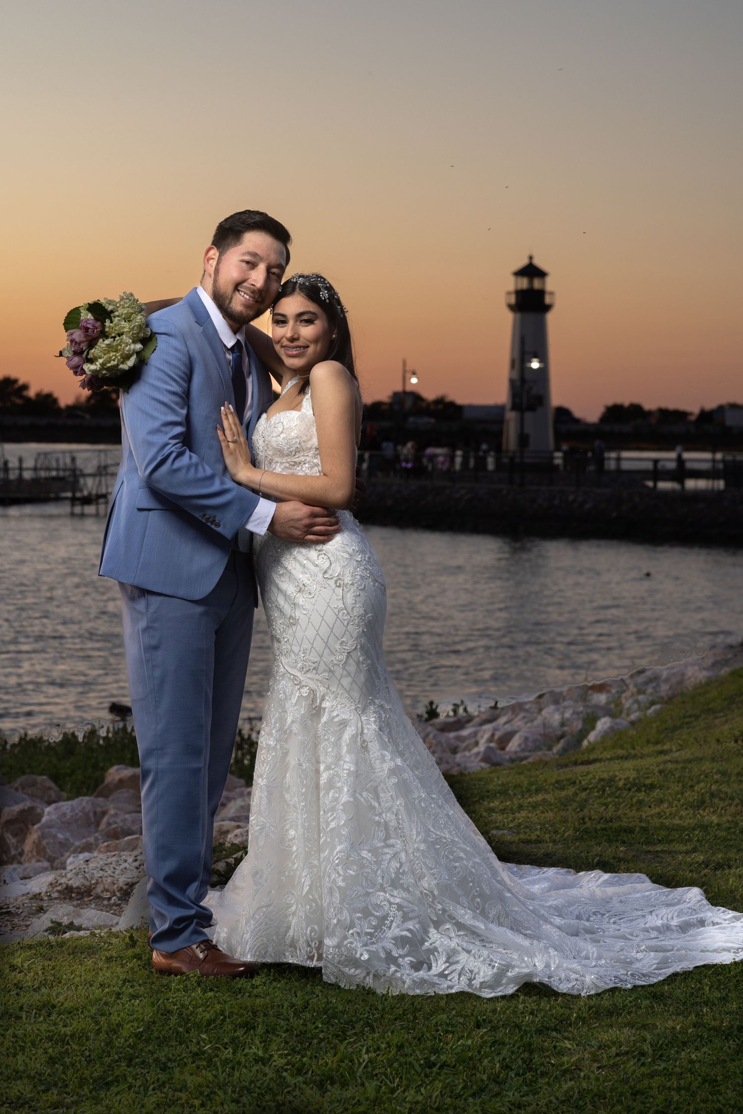 Smiling couple embraces in wedding attire at sunset with lighthouse and waterfront in background.