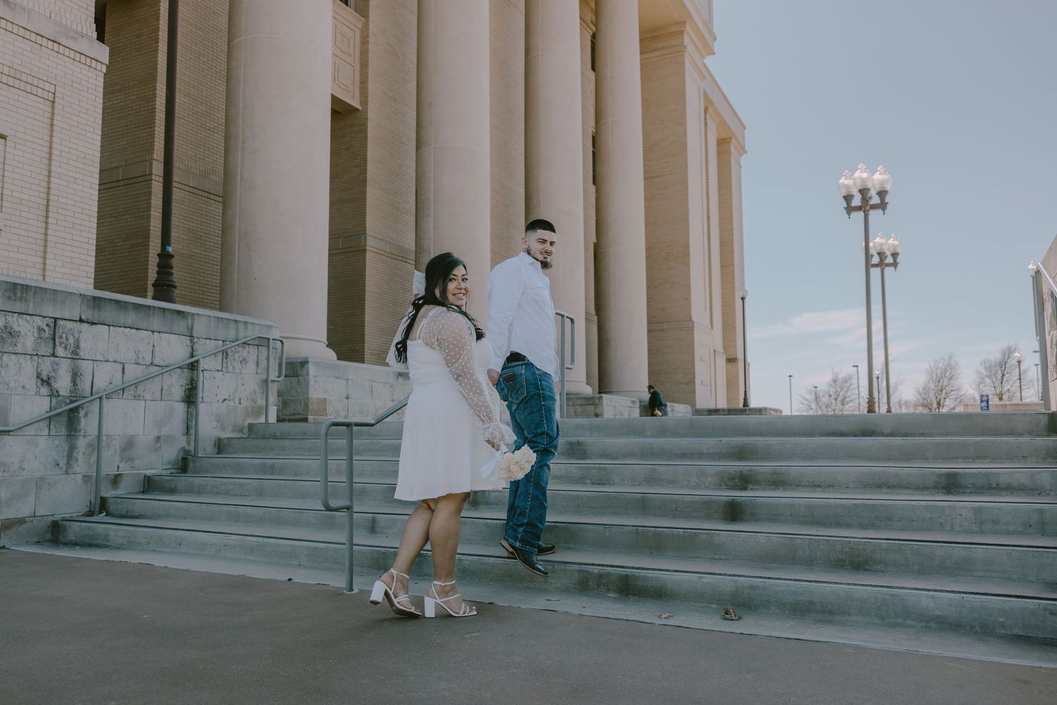 Couple holding hands while walking up grand stone steps of a classical building at dusk.