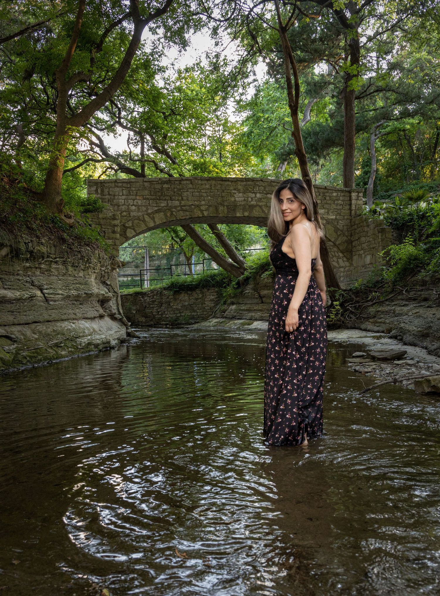 Woman in floral dress standing in creek beneath stone bridge in lush park.