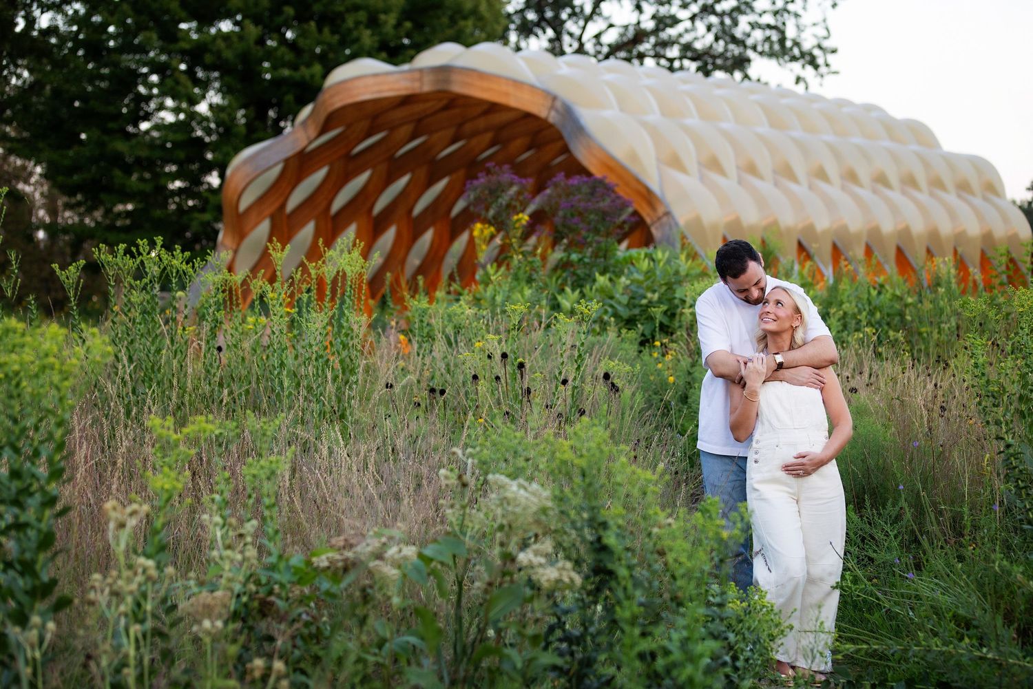 Pregnant couple embracing in lush garden with unique honeycomb wooden structure backdrop during maternity photoshoot.