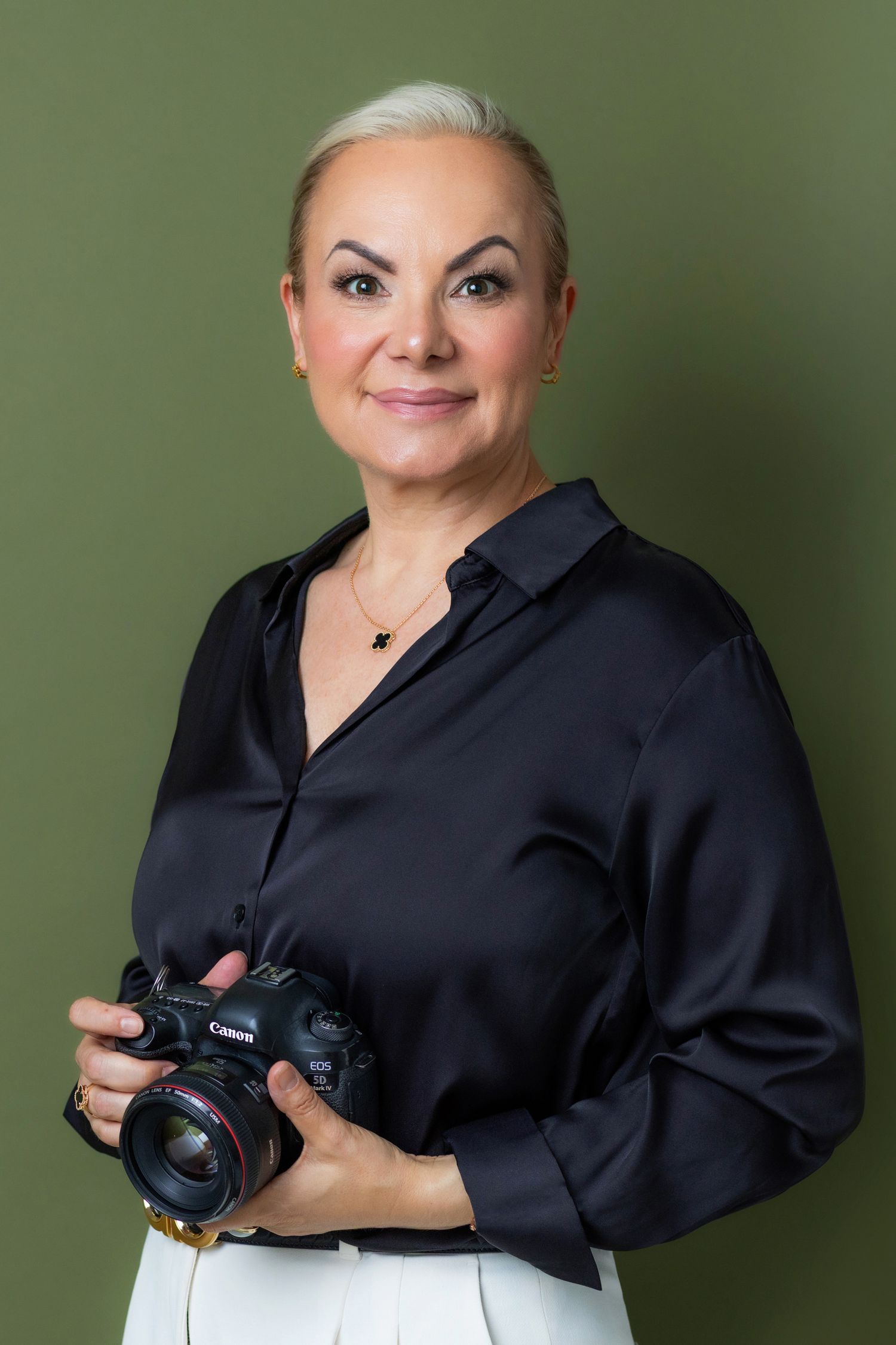 Professional female photographer in black silk blouse holding Canon DSLR camera against olive green background.