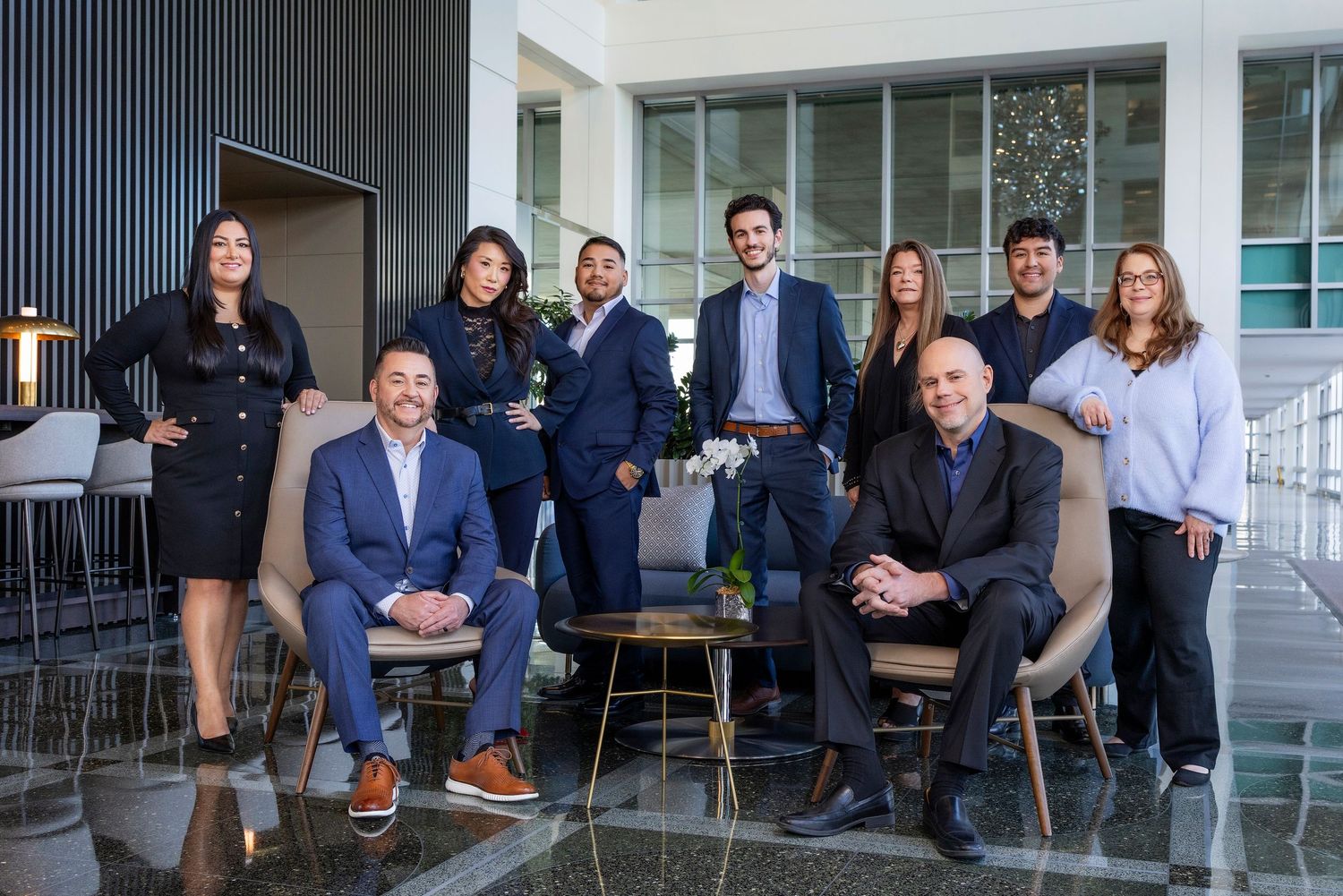 Professional business team members pose together in modern office setting with glass windows and contemporary furniture.