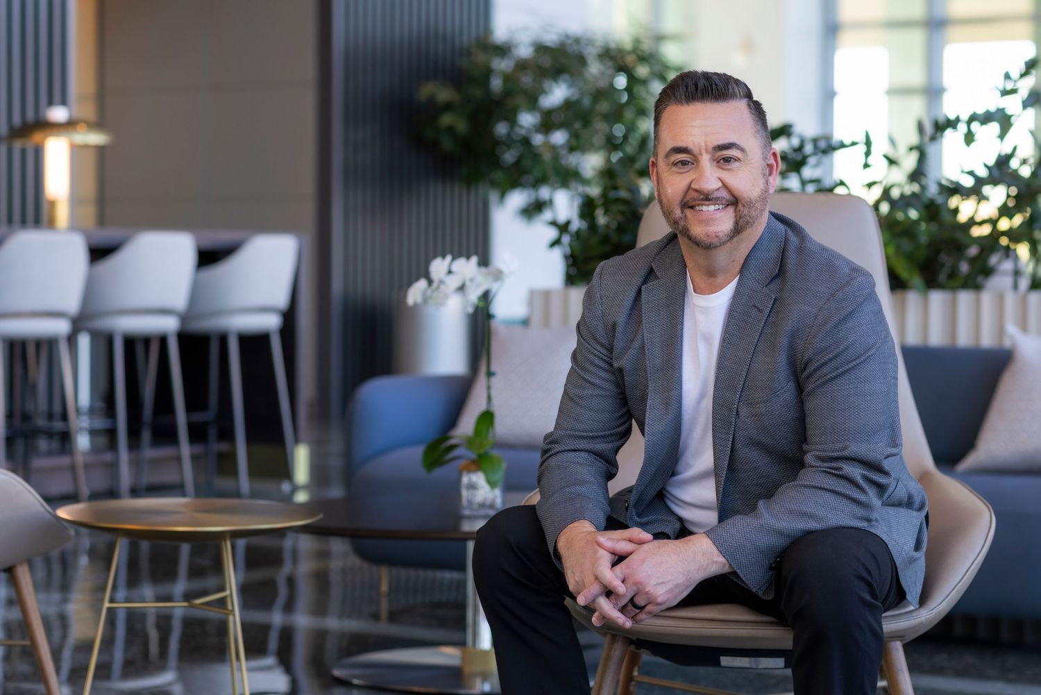 Smiling man in gray blazer sitting in modern office lounge chair with plants and bar stools in background.