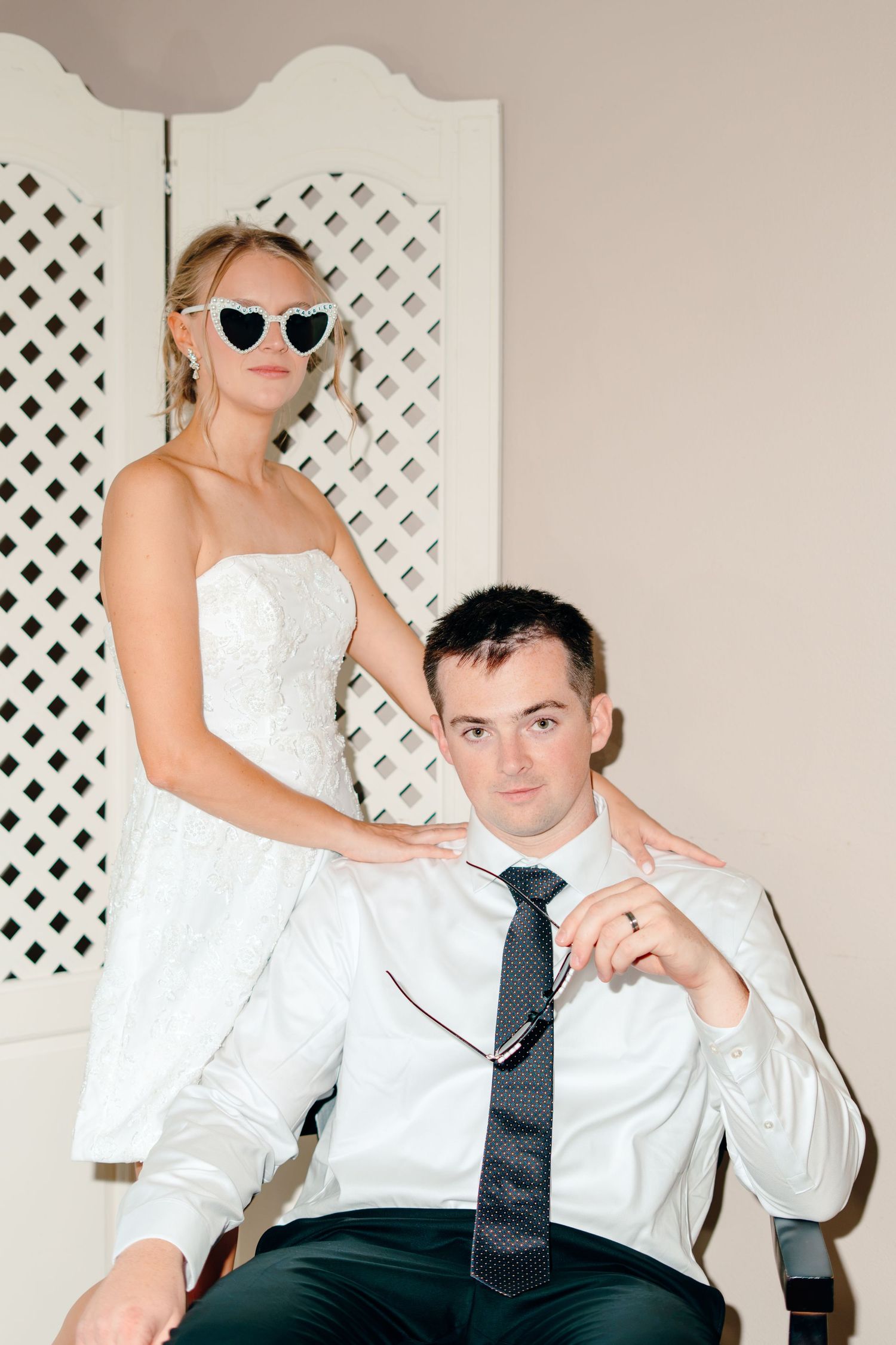 Bride in white dress and heart sunglasses stands behind groom in white shirt and tie seated in chair.