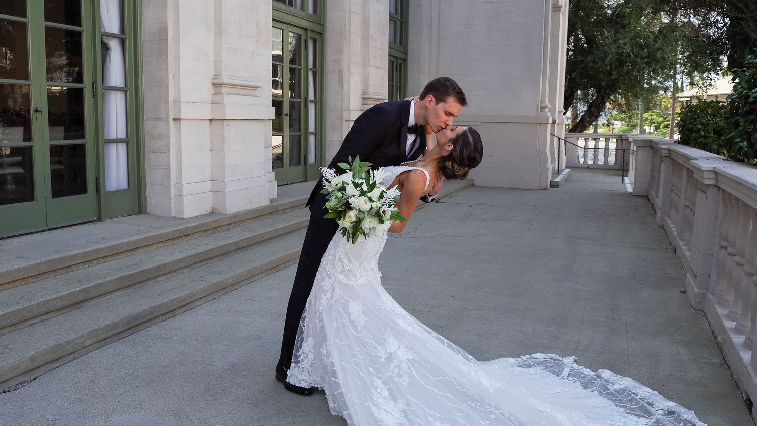 A couple shares a romantic dip kiss on a stone balcony, with the bride in a flowing white gown holding a floral bouquet.