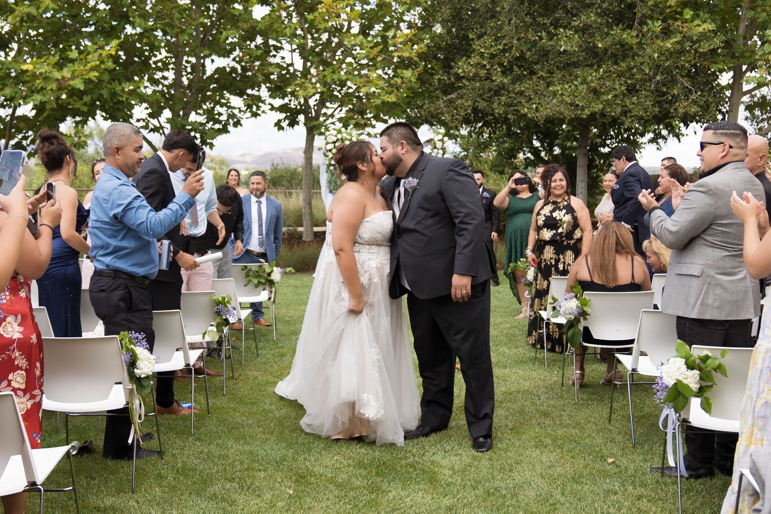 Wedding guests mingle and socialize at an outdoor garden reception with white chairs and lush greenery.