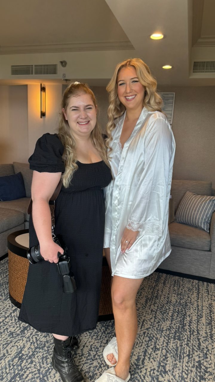Two women pose together indoors, one wearing a black dress and boots, the other in a white button-down shirt dress.