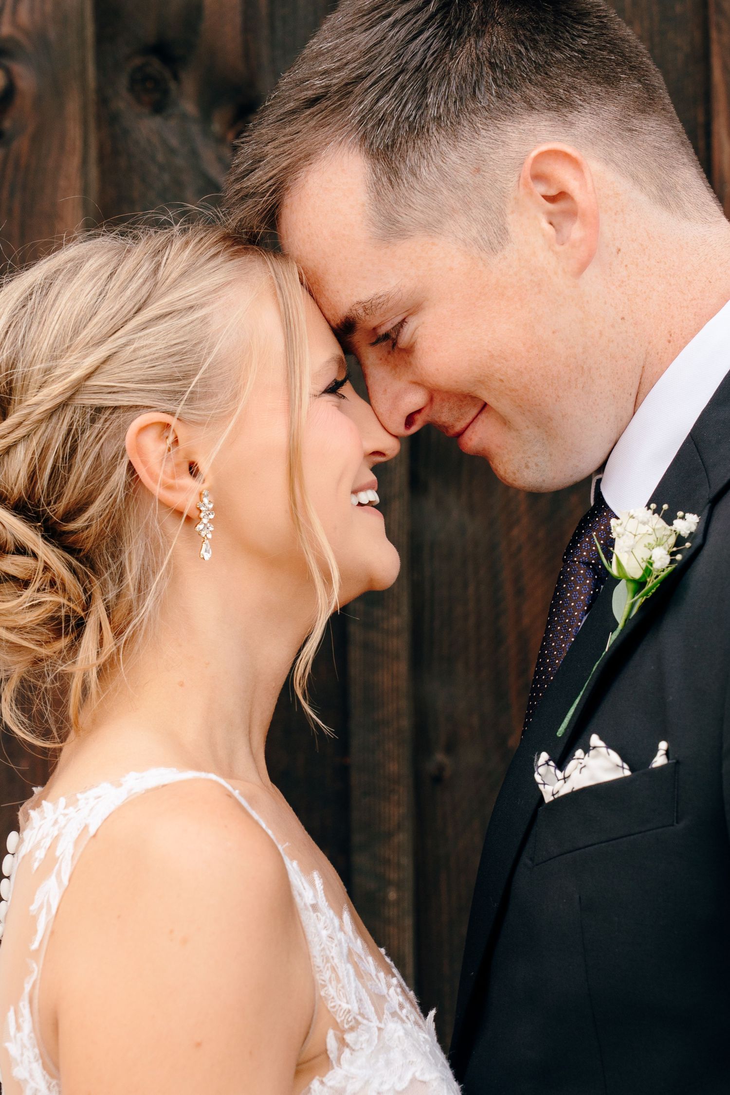 A bride and groom share an intimate moment, touching foreheads and smiling, dressed in wedding attire against a wooden backdrop.