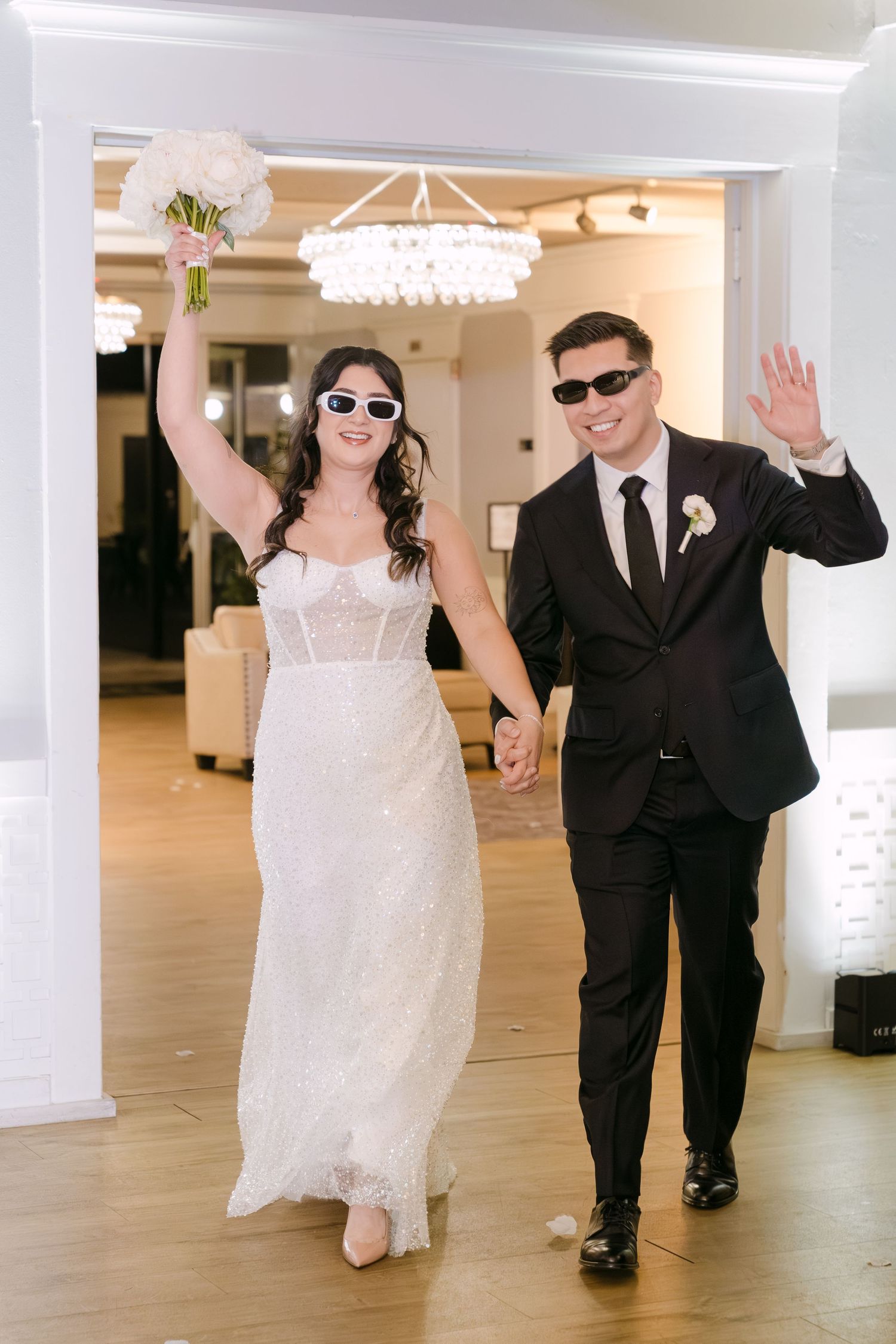 A bride and groom dance and wave while walking through an elegant white-walled venue hallway.