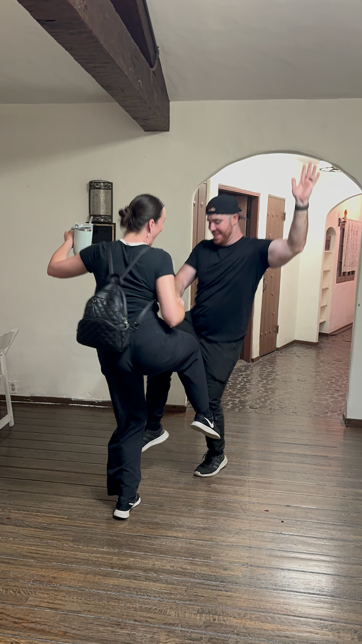Two people dressed in black clothing dance energetically together on a hardwood floor in an indoor space.