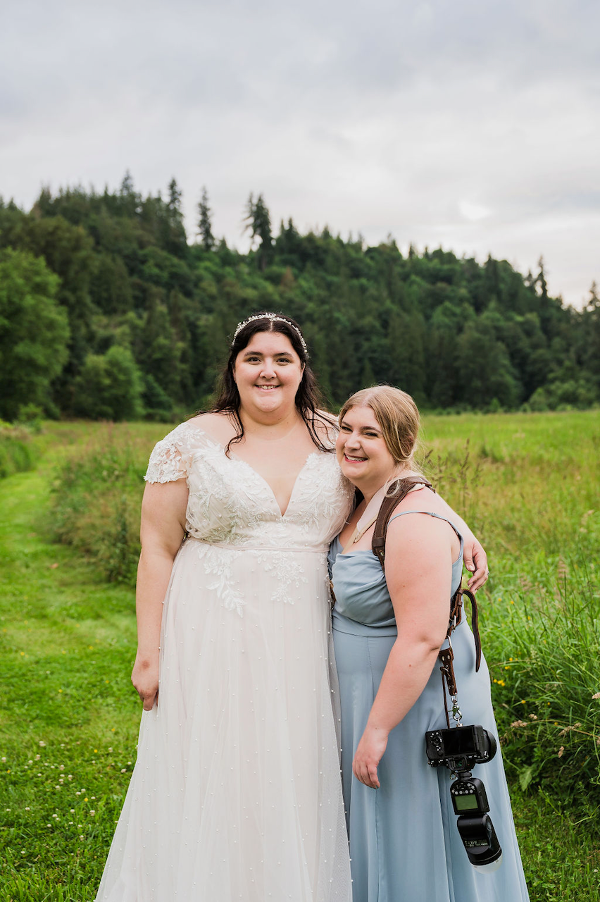 Two people wearing a white wedding dress and blue bridesmaid dress pose together in a green meadow with forest backdrop.