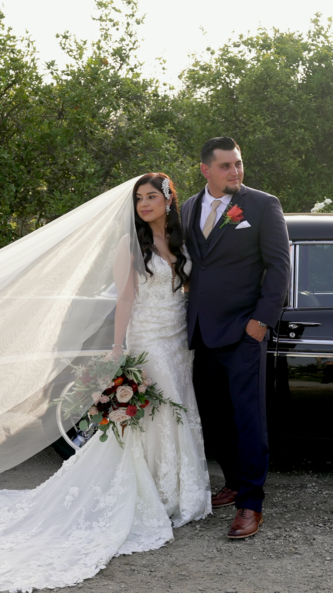 A bride and groom pose together outdoors, with her flowing veil creating a dramatic effect against the natural backdrop.