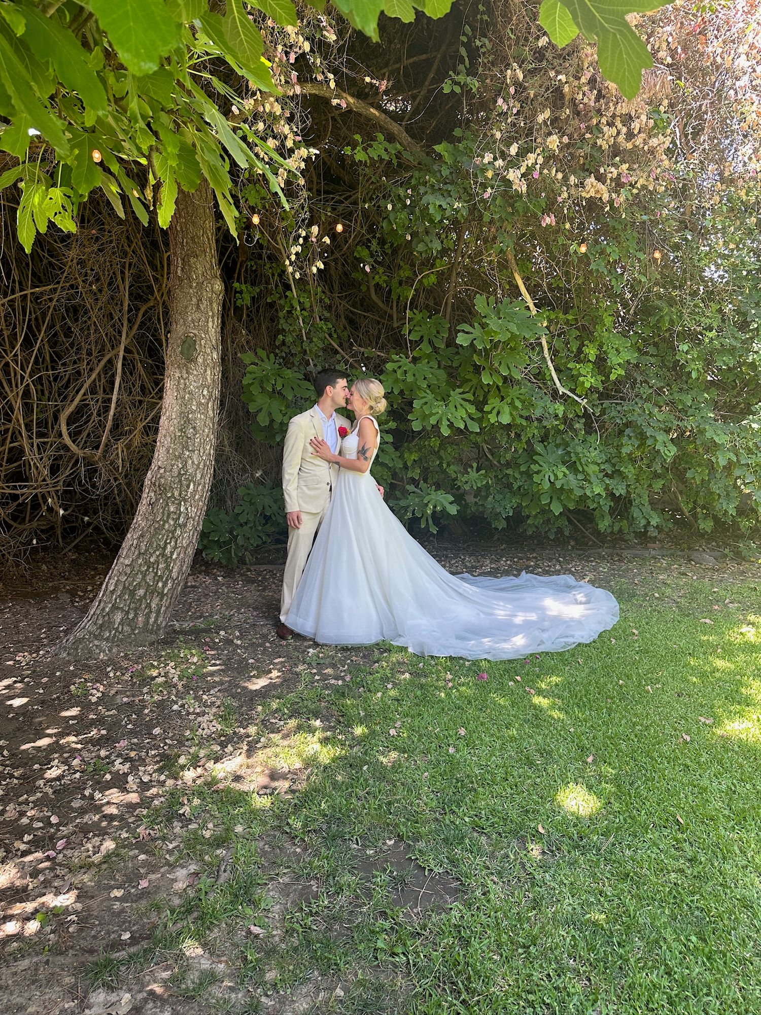 A couple in wedding attire shares an intimate moment under lush green trees in a garden setting.