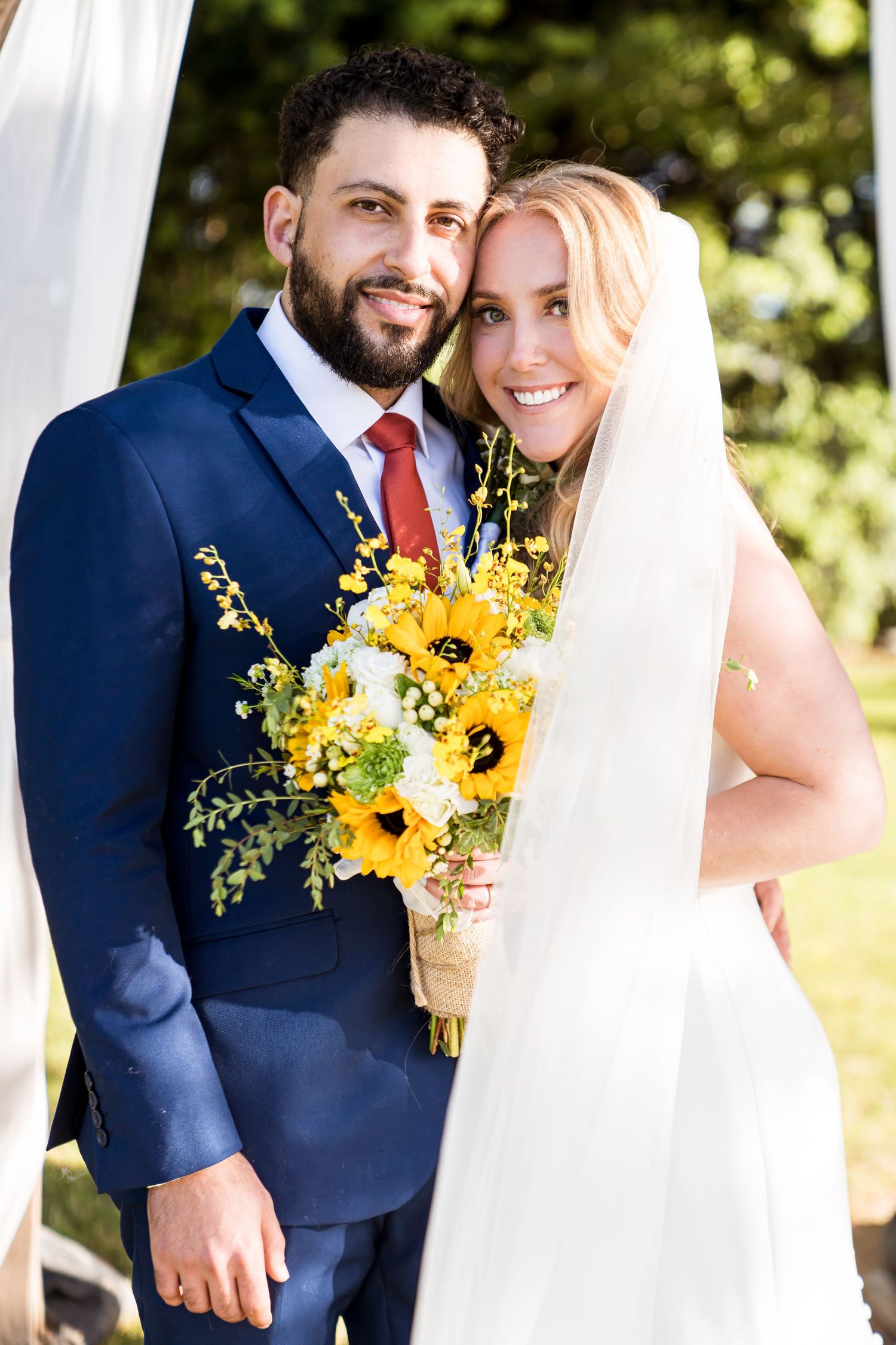 Newlywed couple in navy suit and white dress pose outdoors with bright yellow sunflower bouquet on sunny day.