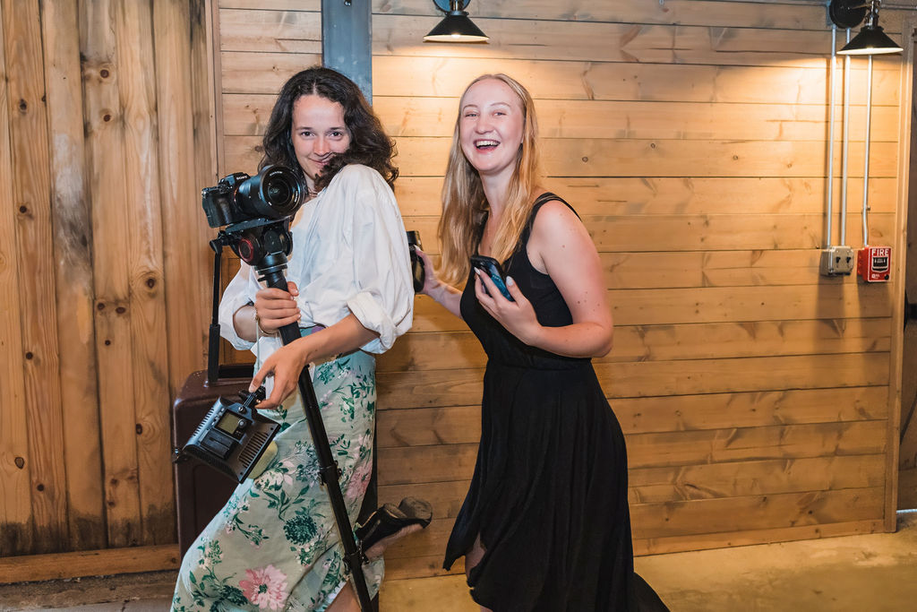 Two photographers with professional cameras pose playfully against a wooden wall with pendant lighting.