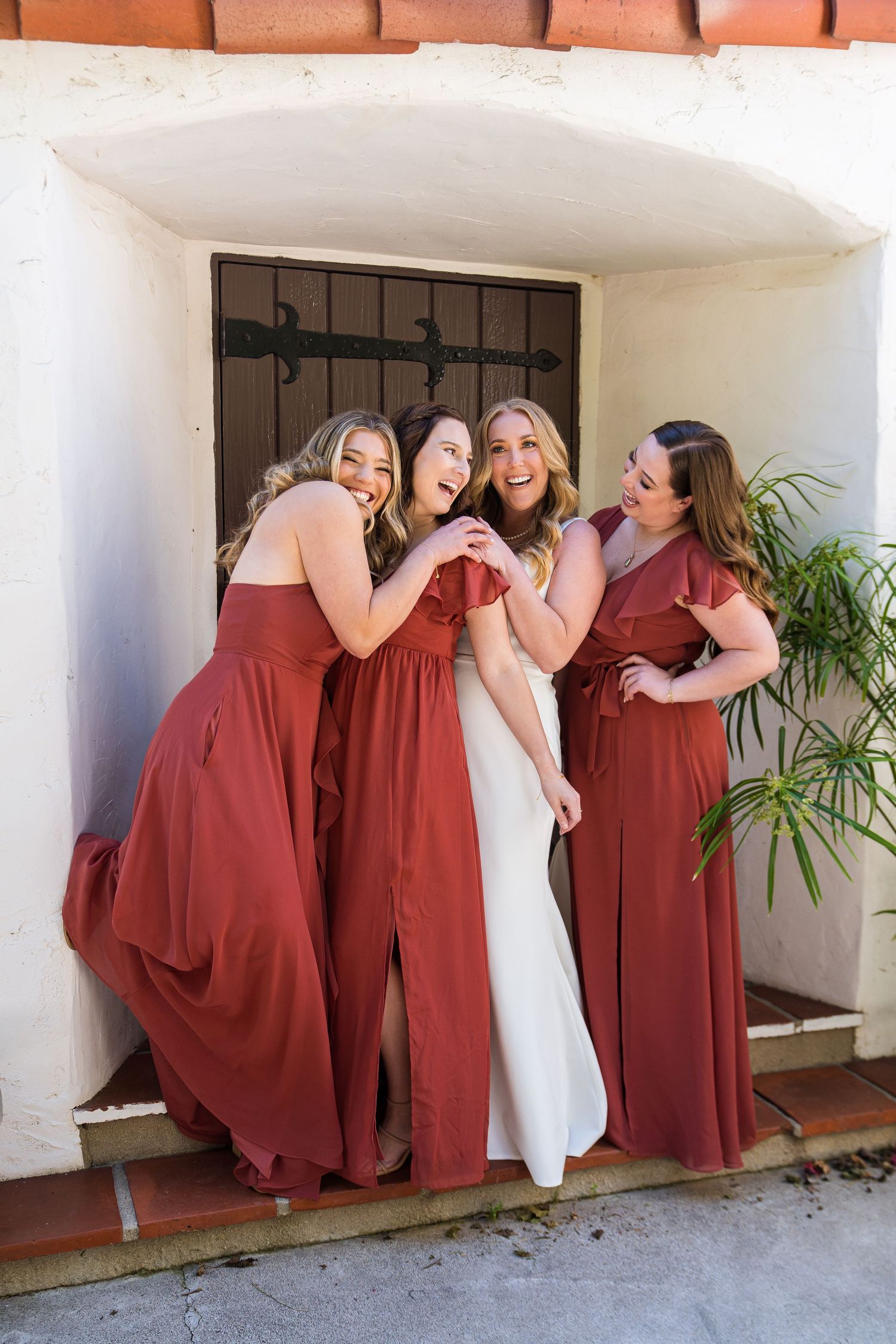 Fun wedding party group in burgundy dresses and white gown pose playfully along outdoor corridor wall.