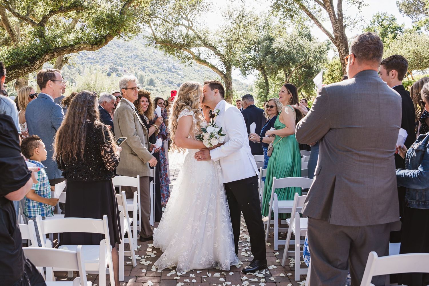 Outdoor wedding ceremony guests mingle and socialize under large trees during a daytime celebration.