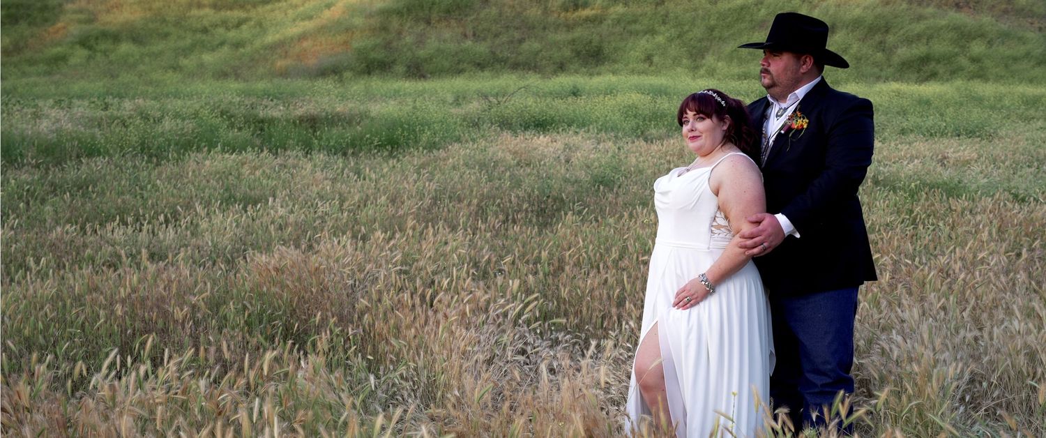 A couple in wedding attire walks through a scenic grassy meadow during sunset.
