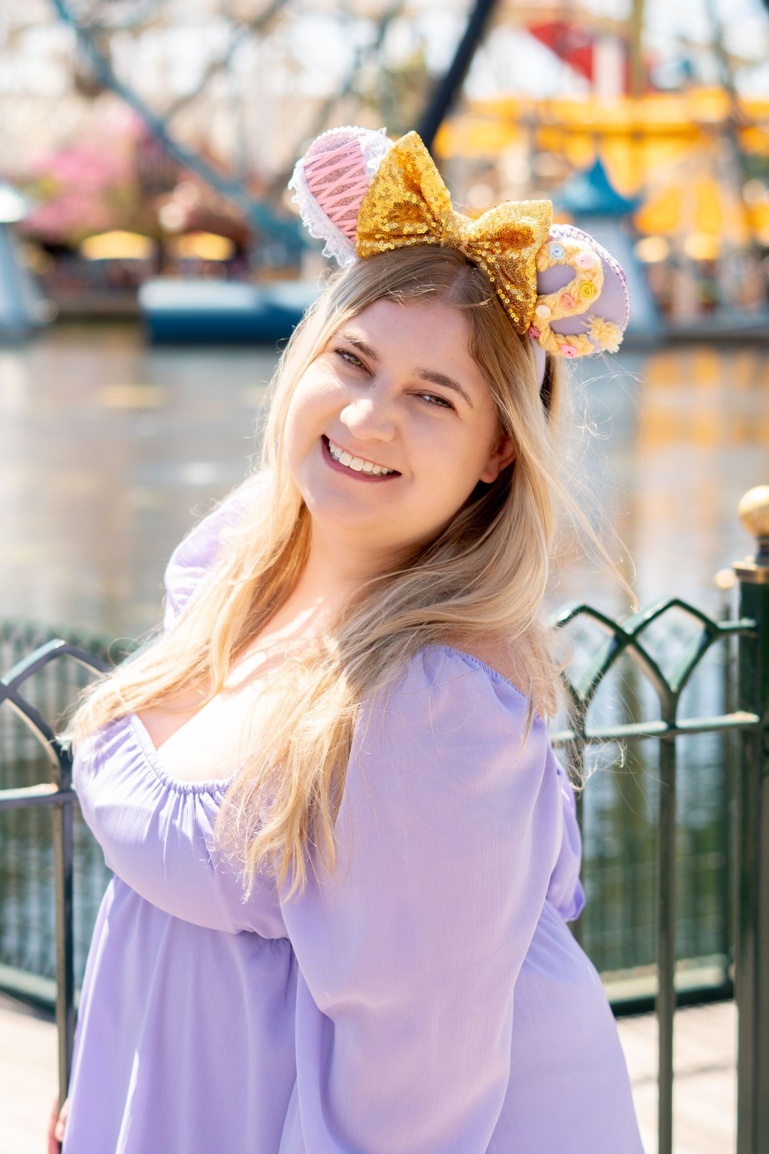 A person wearing a purple dress and gold bow Minnie Mouse ears smiles at a Disney theme park.