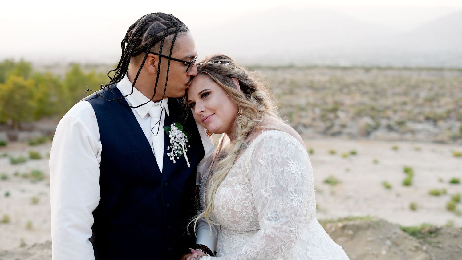 A couple shares an intimate moment in a desert landscape wearing formal wedding attire.