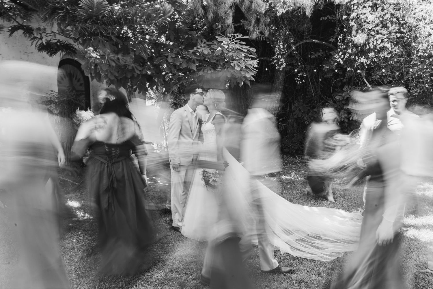 Black and white wedding photo of couple kissing surrounded by blurred moving guests outdoors.