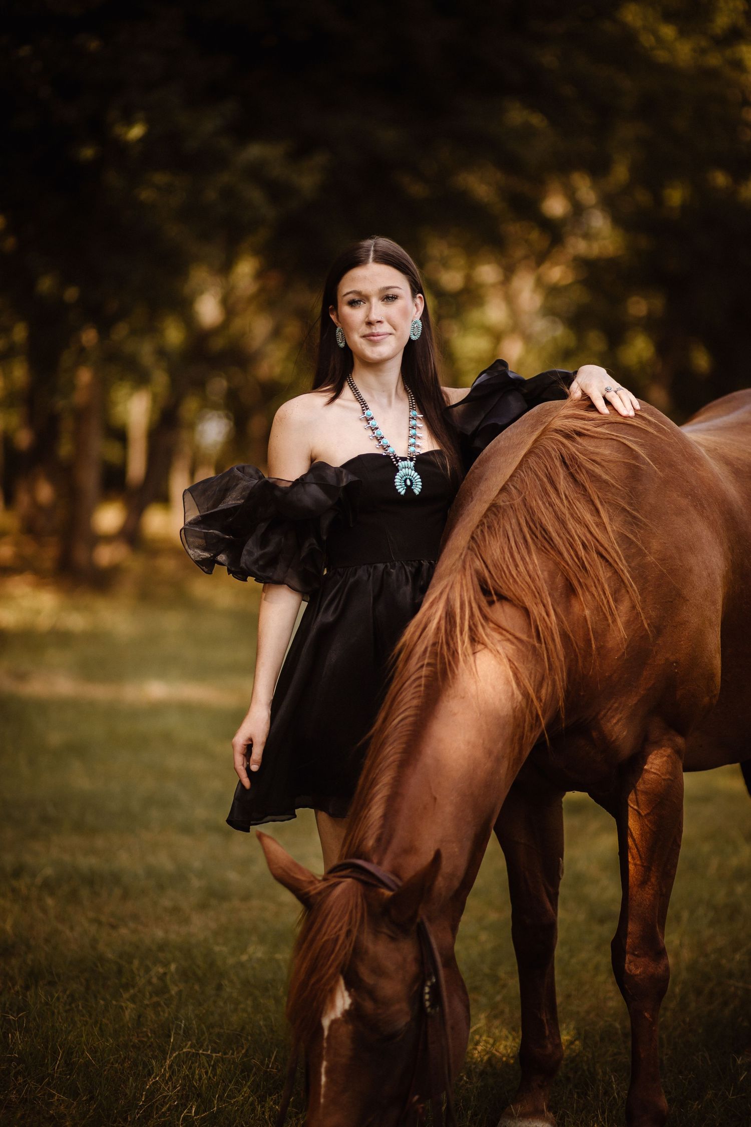 Person in black dress poses with brown horse in sunlit field.