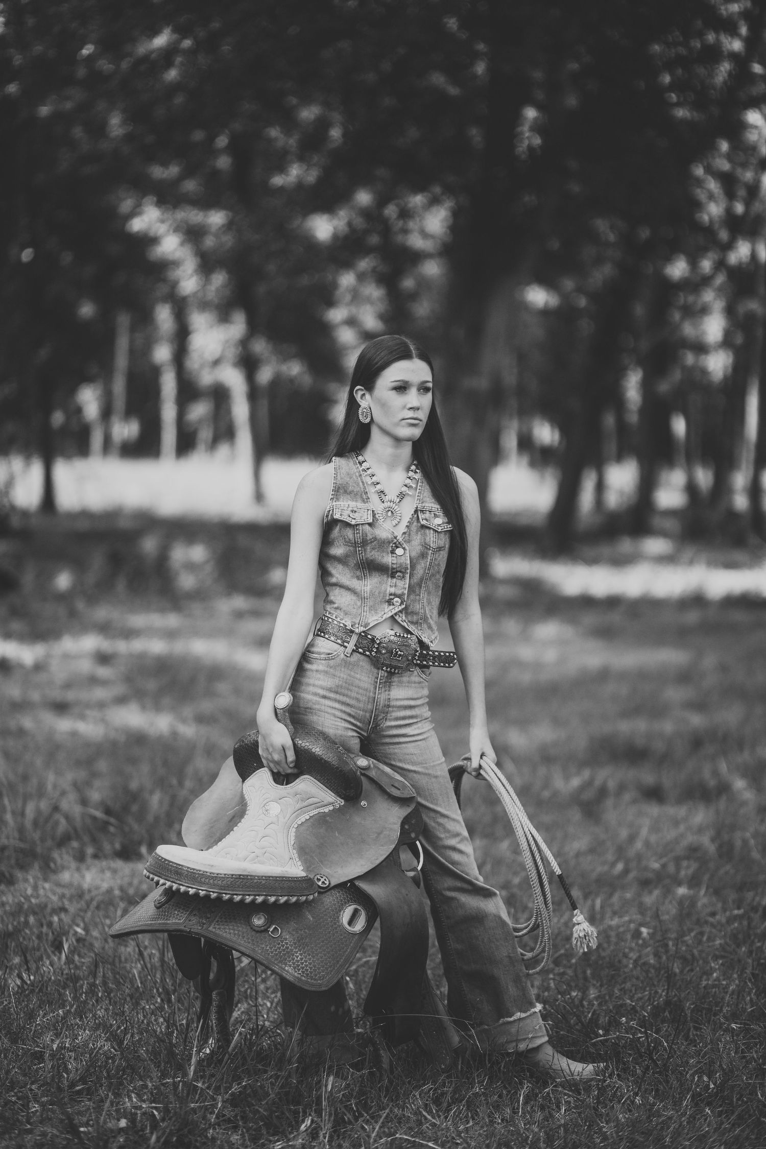 A person in overalls stands with a rustic wooden saddle in a grassy field with trees in the background.