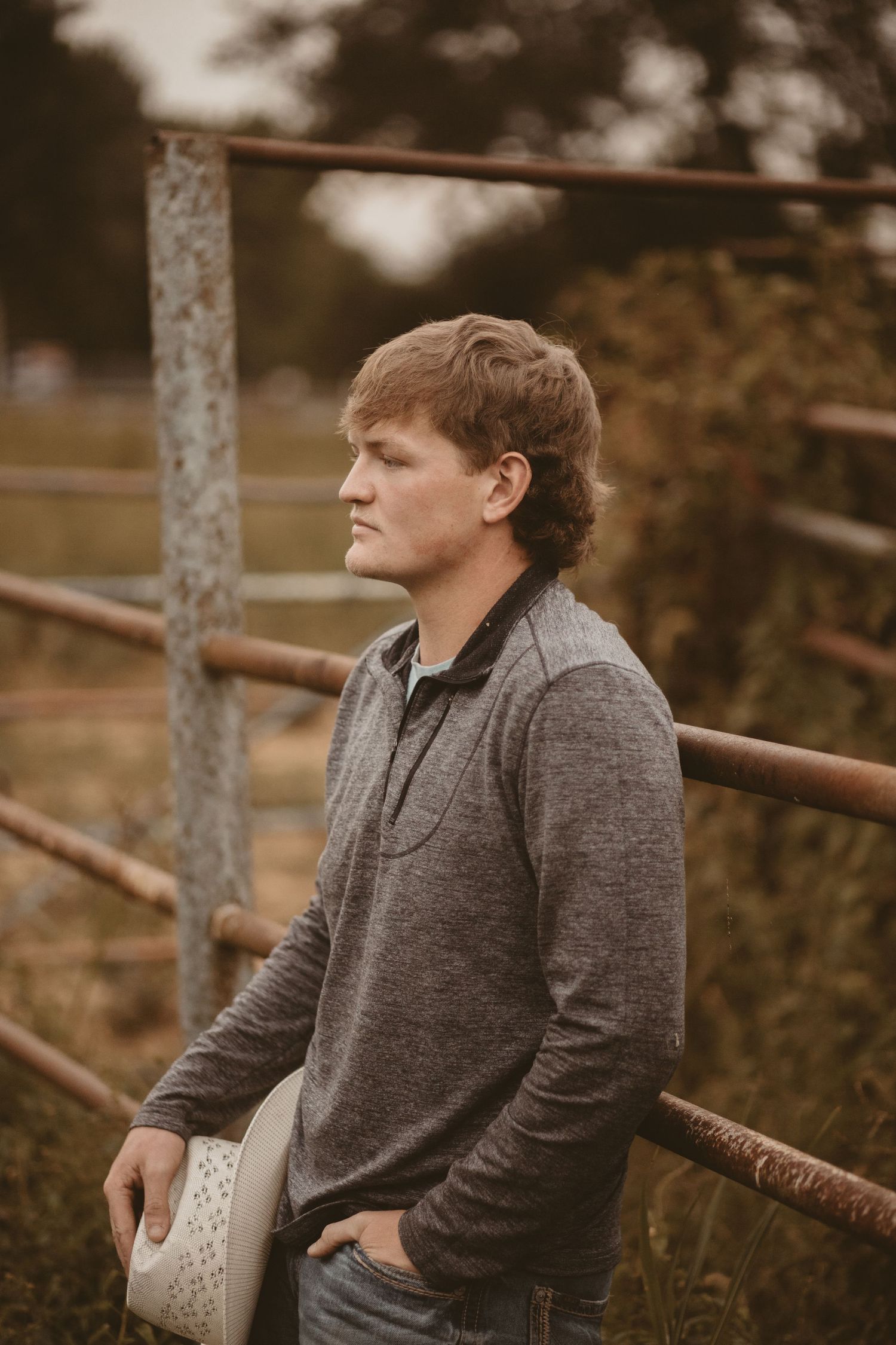 Person in a grey sweater leans against wooden fence posts in warm autumn lighting at the old rodeo arena with a cowboy hat.