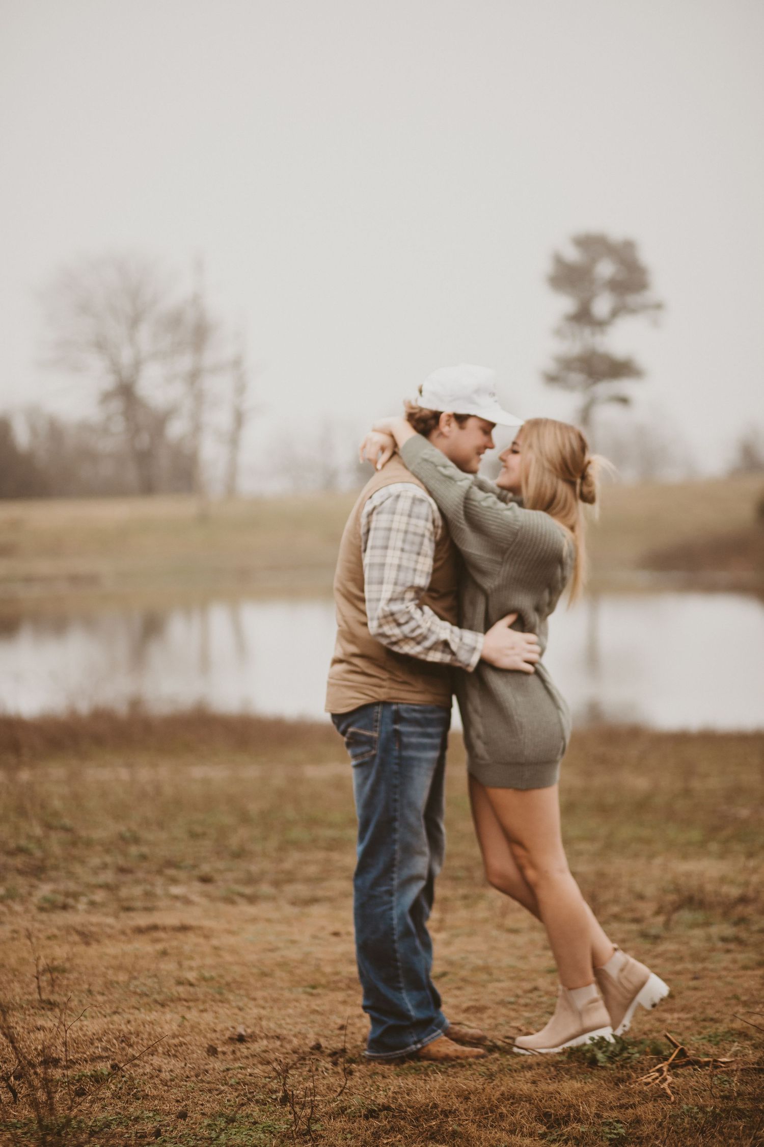 Romantic couple embraces in a rustic outdoor setting near a pond at sunset.