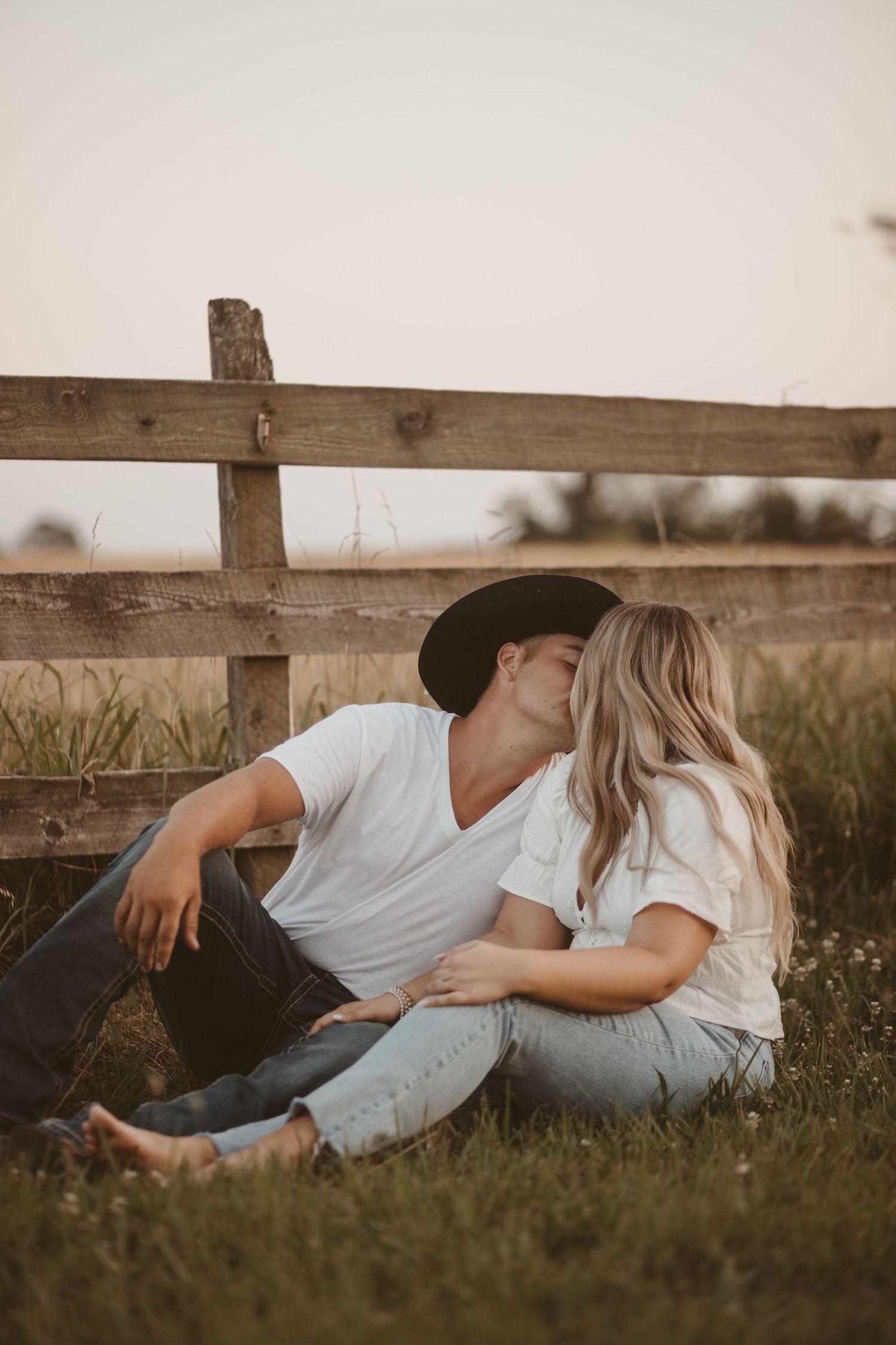 Couple relaxes by wooden fence in grass field during golden hour.