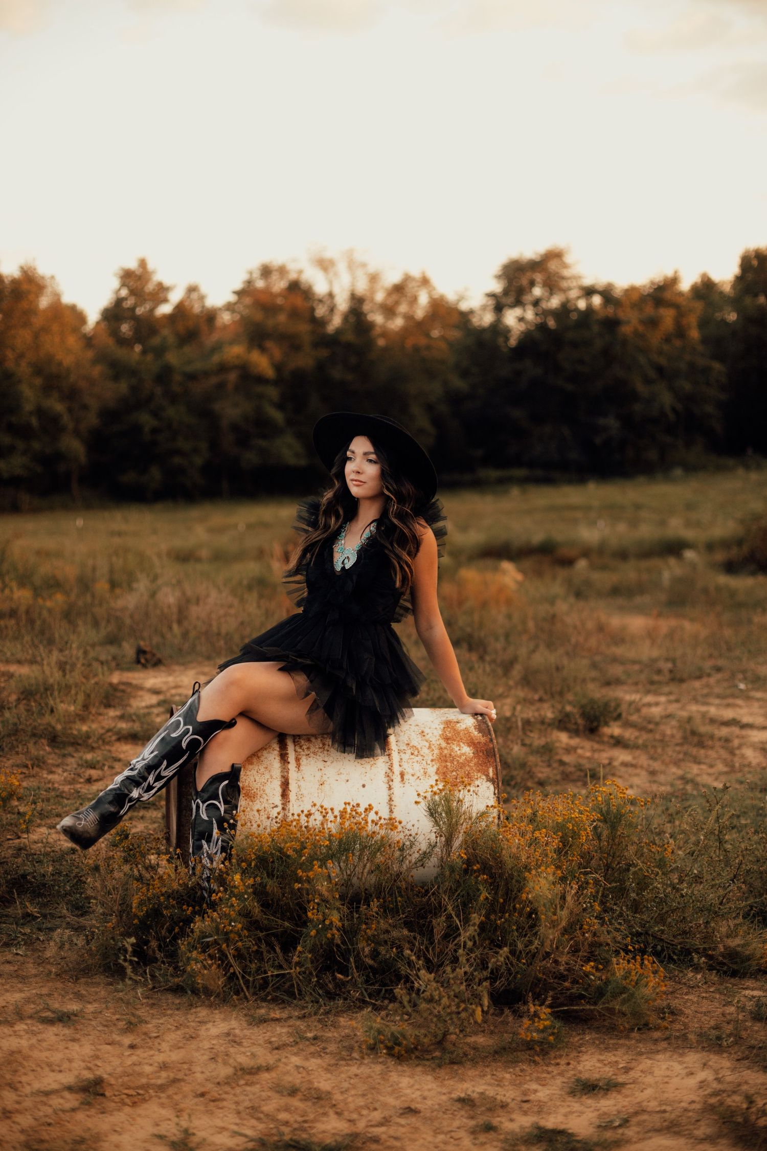 Artistic outdoor cowgirl photography session in a field at golden hour with flowing black lace dress and boots.