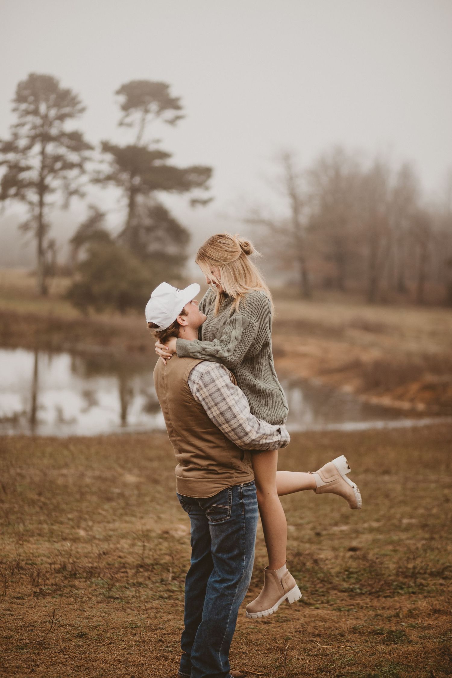 A couple embraces in a field with a lake and trees visible in the muted autumn landscape.