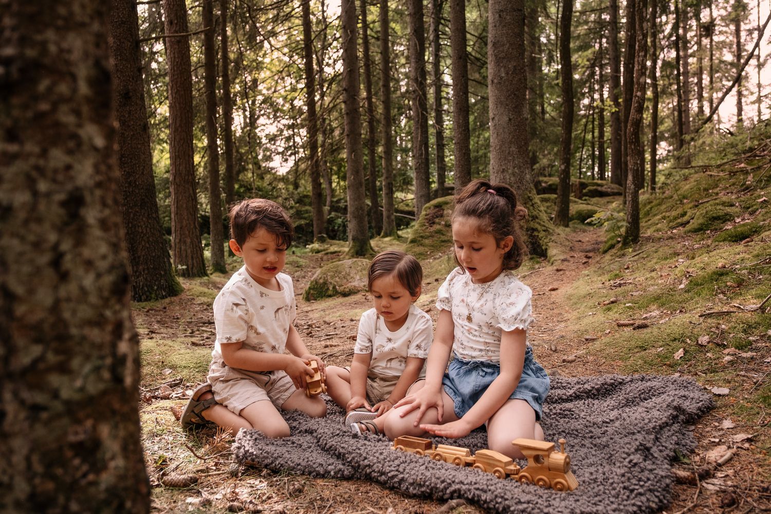 Tre syskon sitter tillsammans på en filt och leker med trätåg i en skogsglänta.