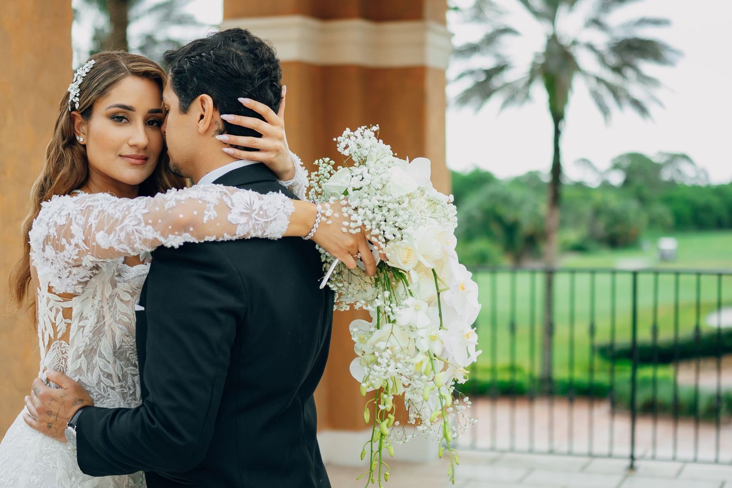 A couple embraces on their wedding day with palm trees and greenery in the background.
