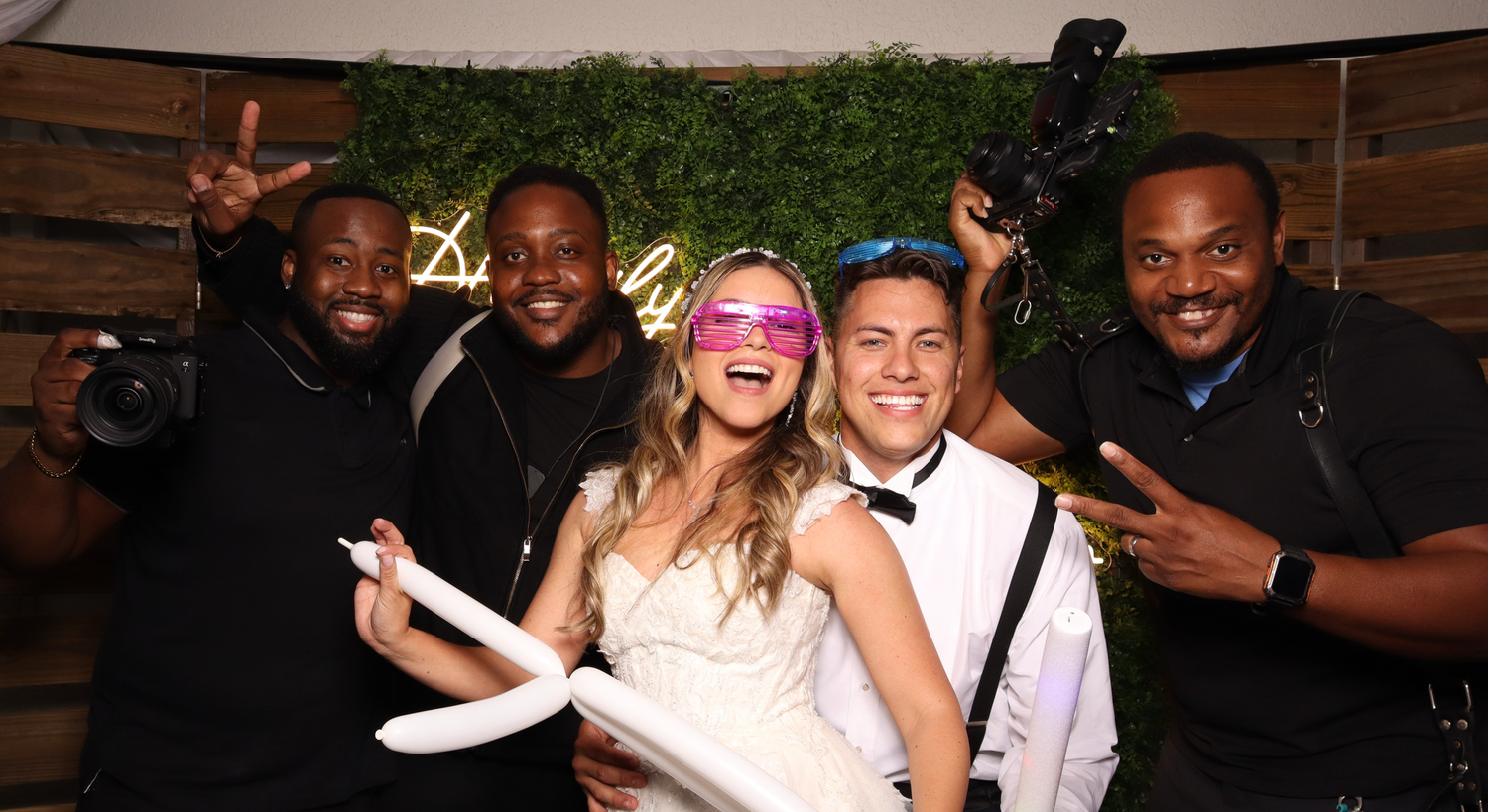 Five friends posing together at a party with decorative lighting and greenery backdrop.