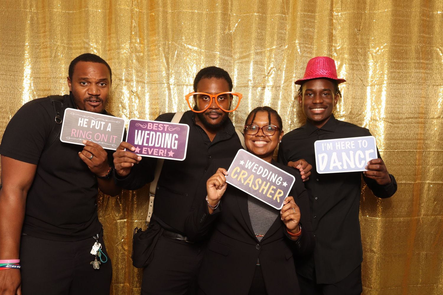 Four people pose with fun photo booth signs against a gold sequin backdrop at a wedding celebration.