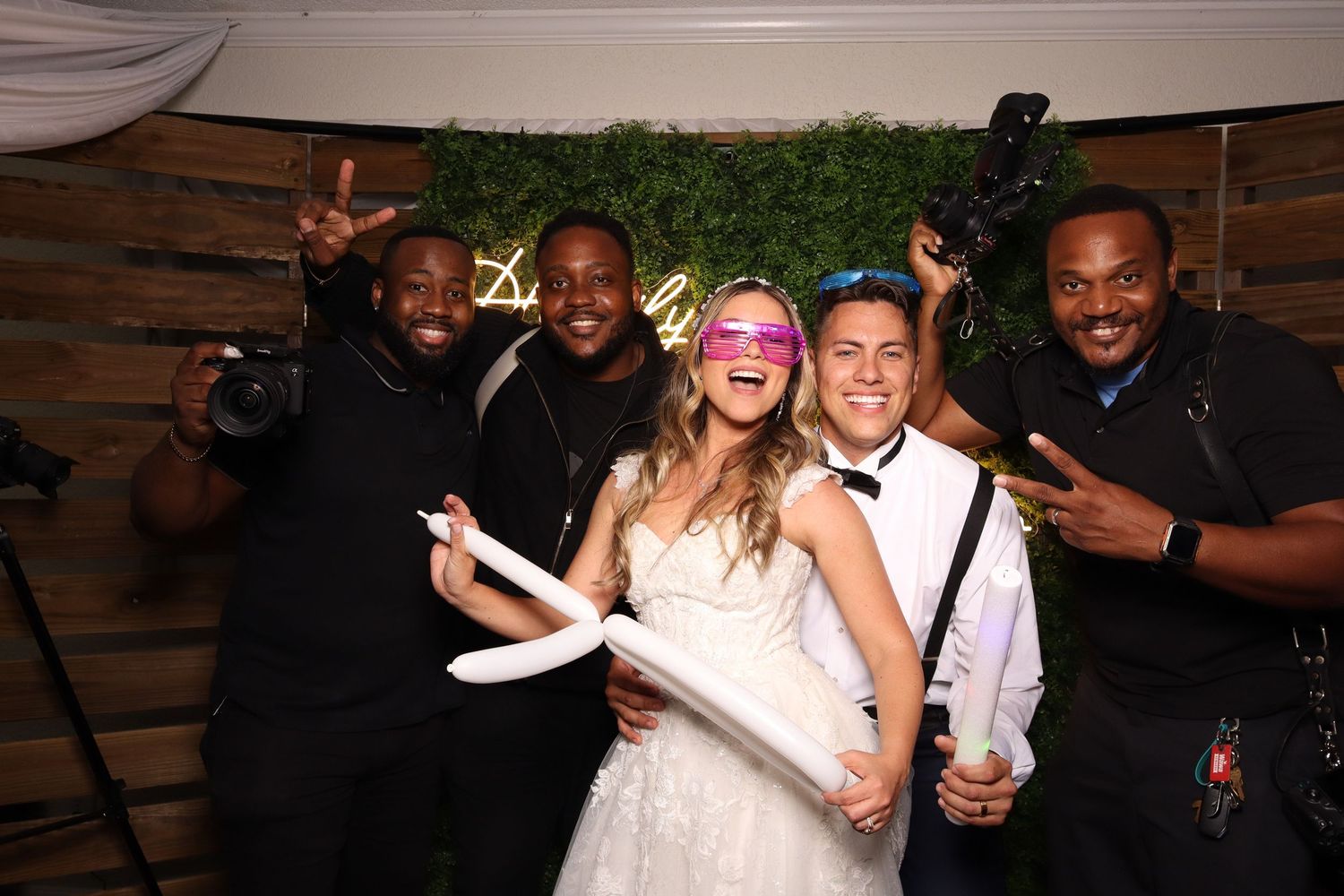 Wedding party celebrates at photo booth with bride and guests posing together against rustic wooden backdrop.