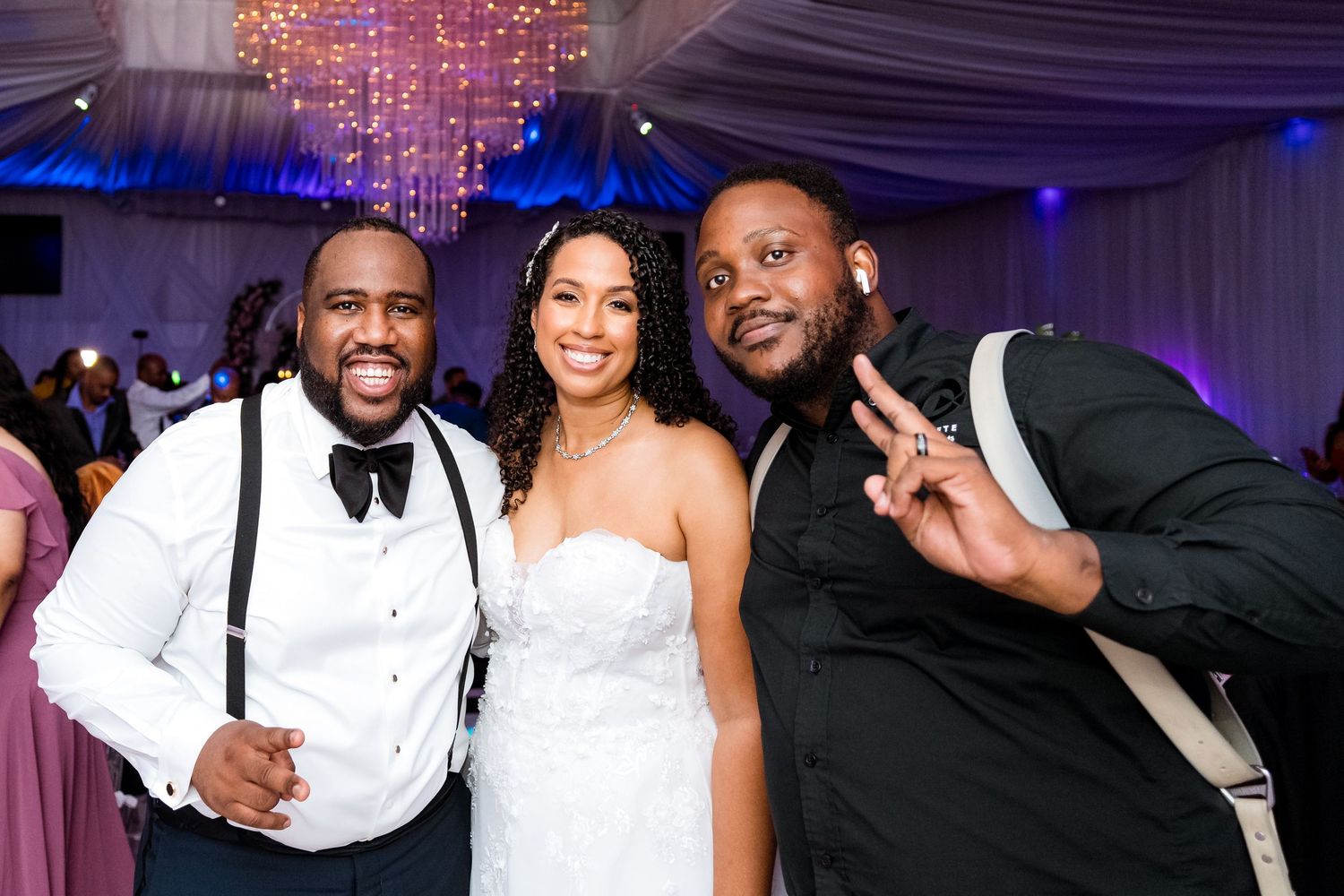 Three guests pose together at an elegant wedding reception with purple lighting and crystal chandeliers in the background.