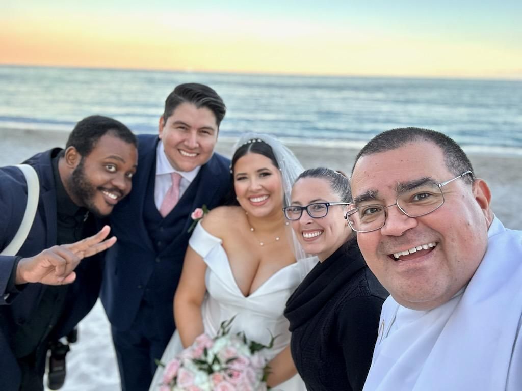 A group of friends pose together for a selfie at a beach during sunset with a bride in white dress holding flowers.